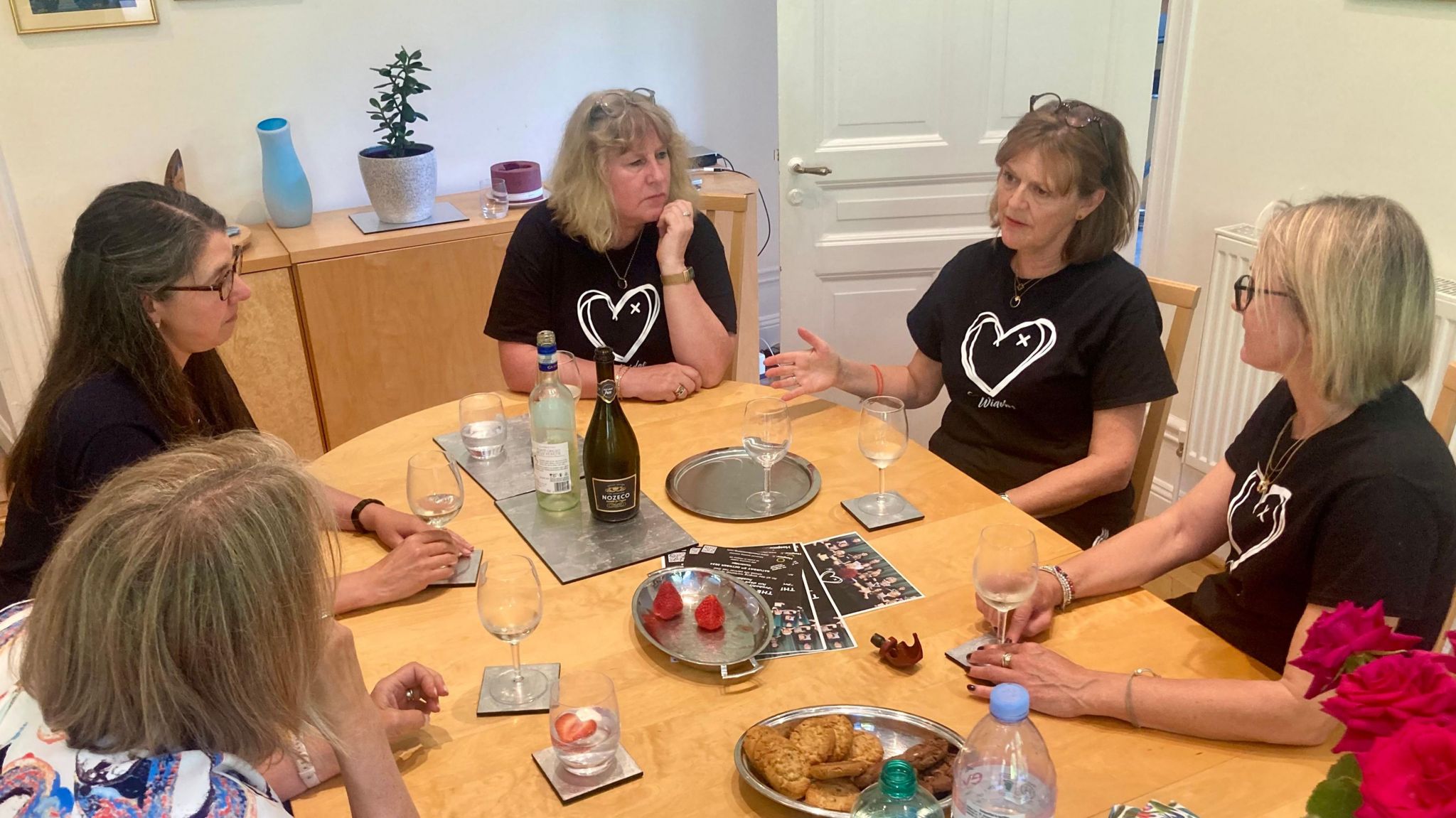 A group of five women sitting at a pine table, which has glasses, two bottles and biscuits in a dish, listening to one of them speaks 