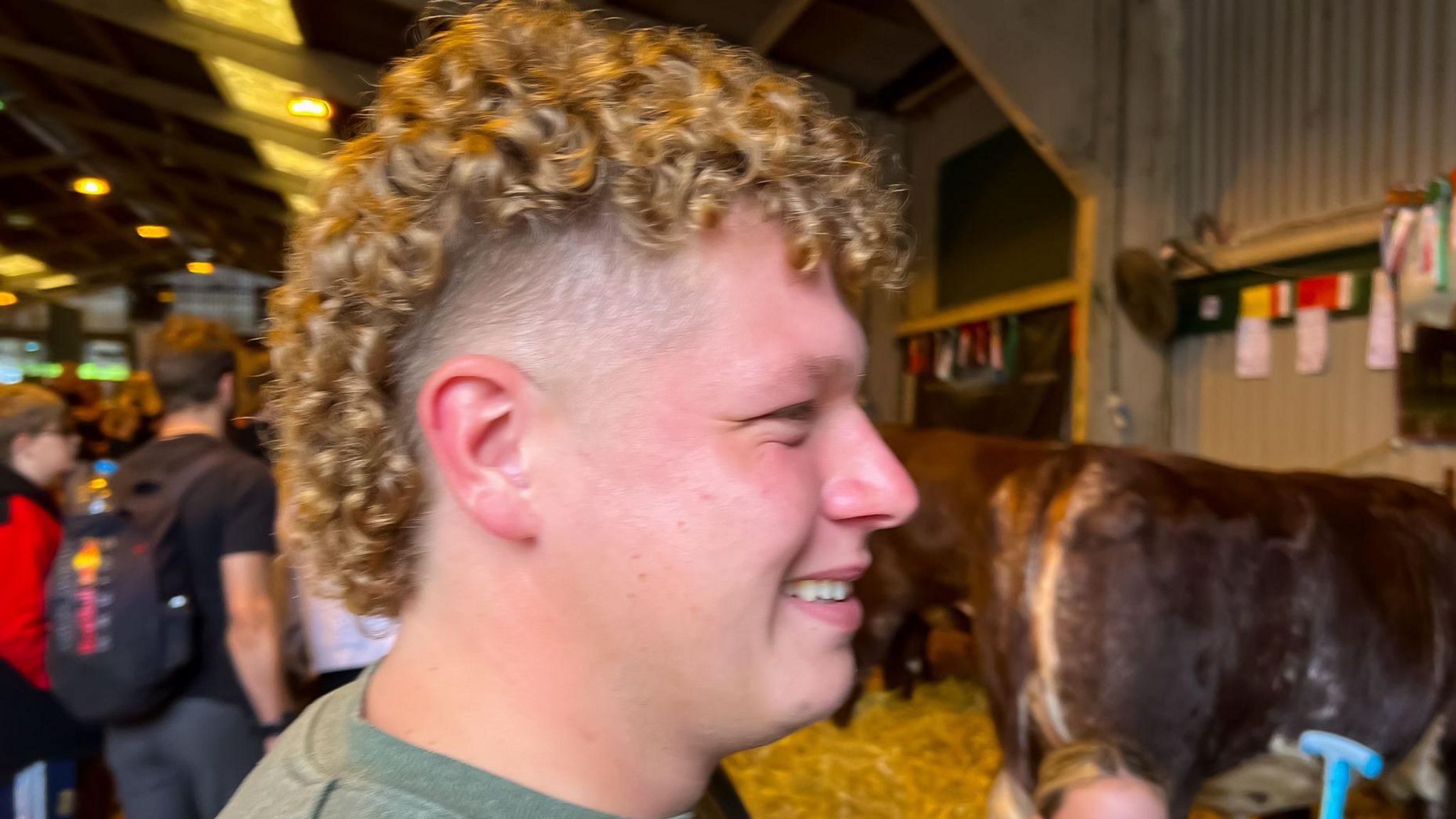 Smiling man in a cow shed with long curly blonde hair on top and in the back and shaved at the sides 