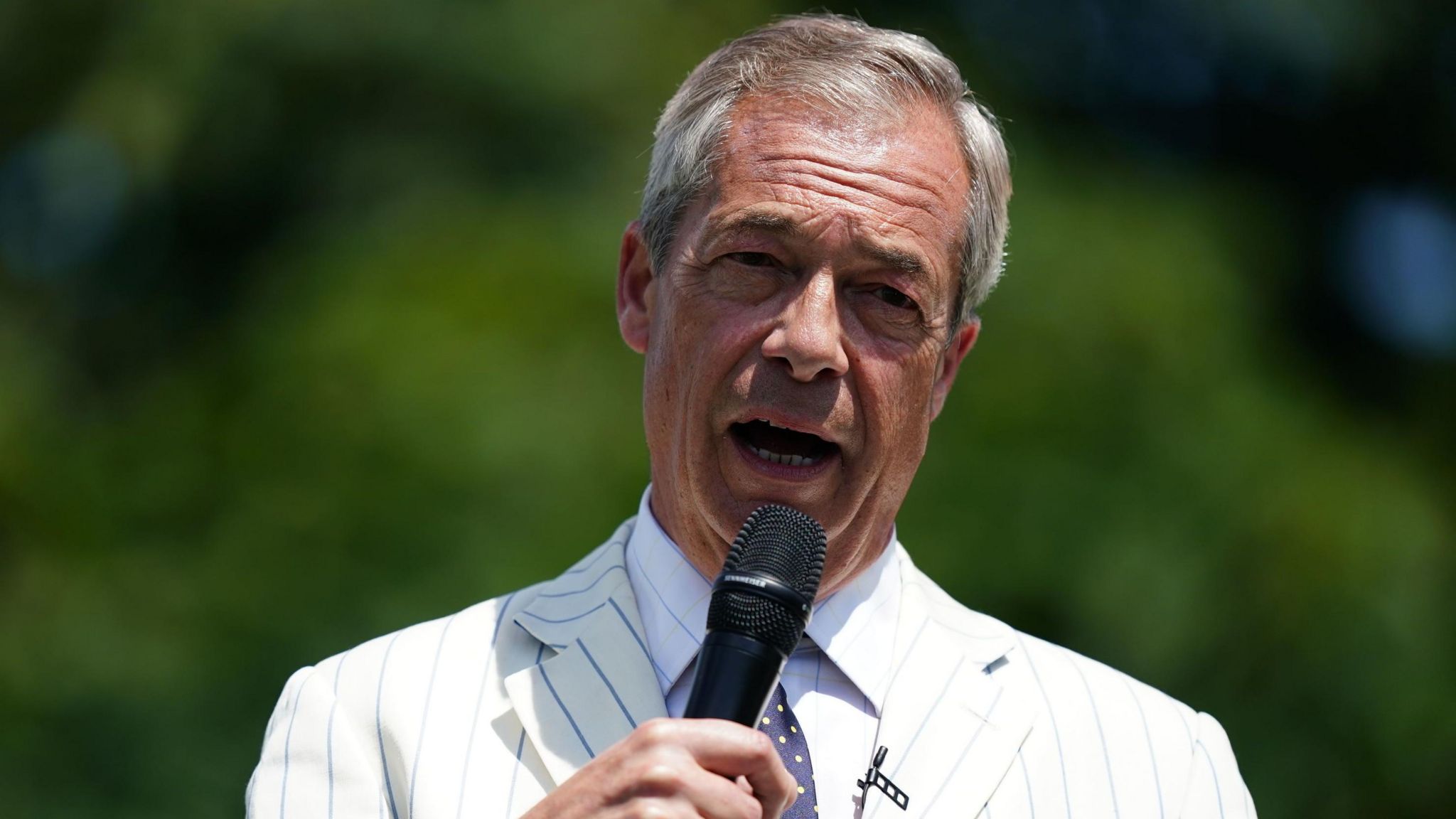 Nigel Farage, wearing a white jacket with blue stripes, speaking into a microphone