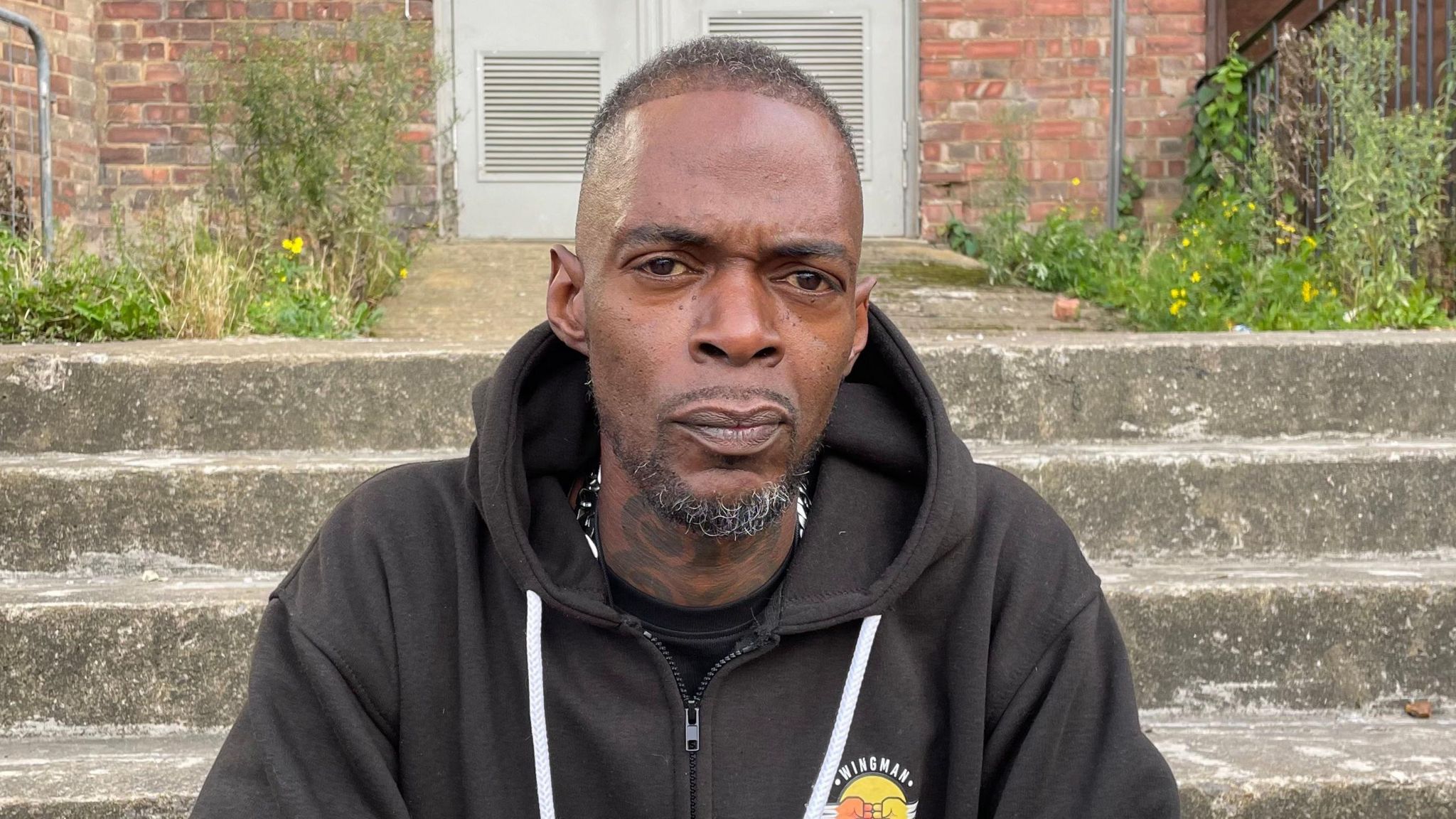 Si Philbert pictured looking direct to camera. He is seated on some concrete steps outdoors. There are steps leading up to a door behind him and weeds growing along the side of a short path between the steps and the door. Mr Philbert is wearing a black hoodie with a white drawstring. He has dark brown eyes and short black hair. He wears a goatee beard, some of whose bristles are turning grey.  