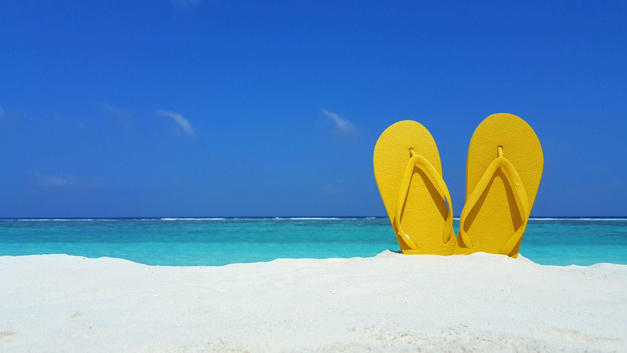 A pair of yellow flip-flops protrude from the sand on a sunny beach, with a blue sky in the background