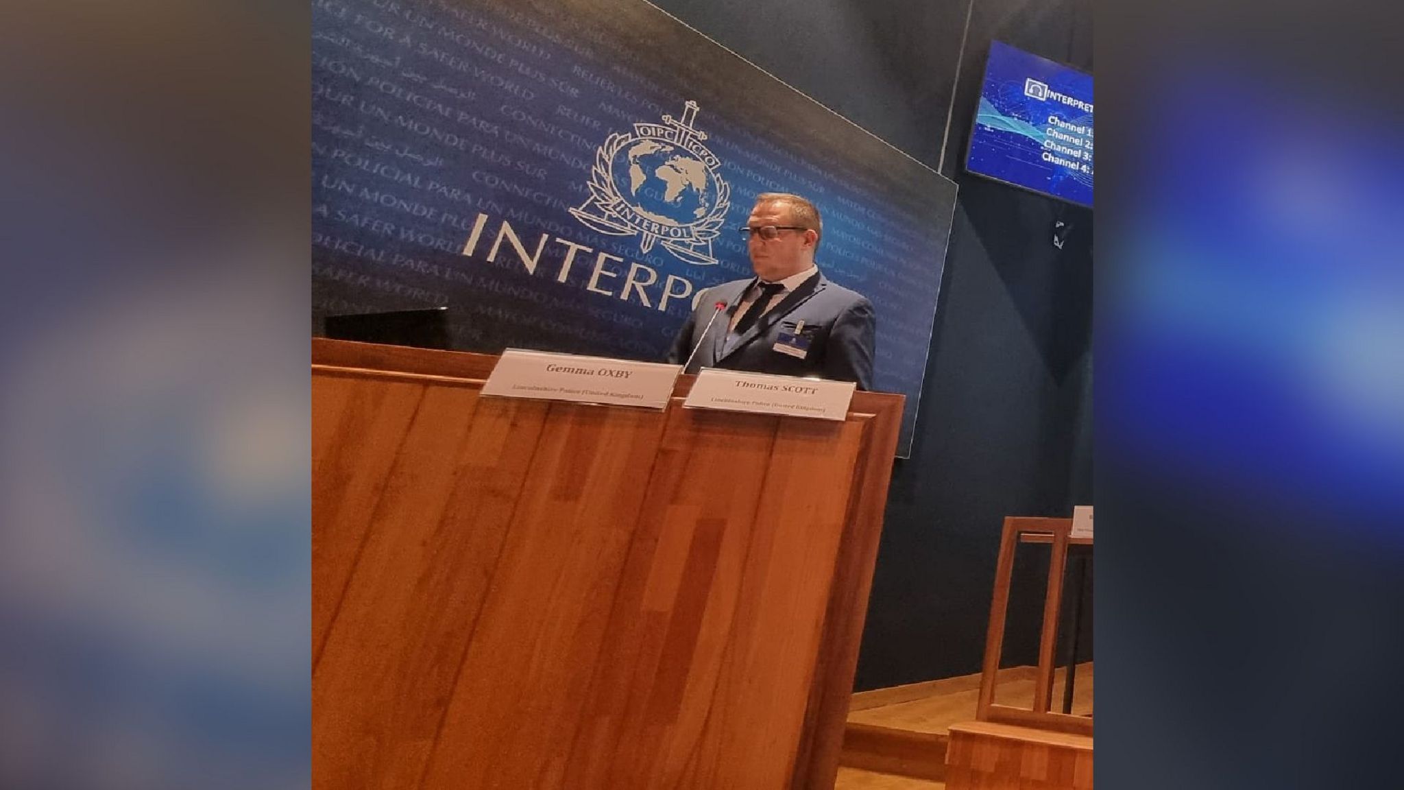 PC Tom Scott, who wears glasses and has short hair, stands in police uniform behind a lectern in a conference hall. The Interpol logo is on a screen behind him. 