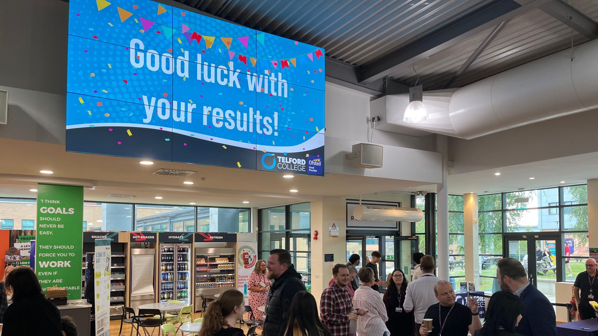 A large television screen is mounted on a wall near the ceiling of a college building. The screen is blue with colourful banners, and reads "good luck with your results!". There are people walking below it.