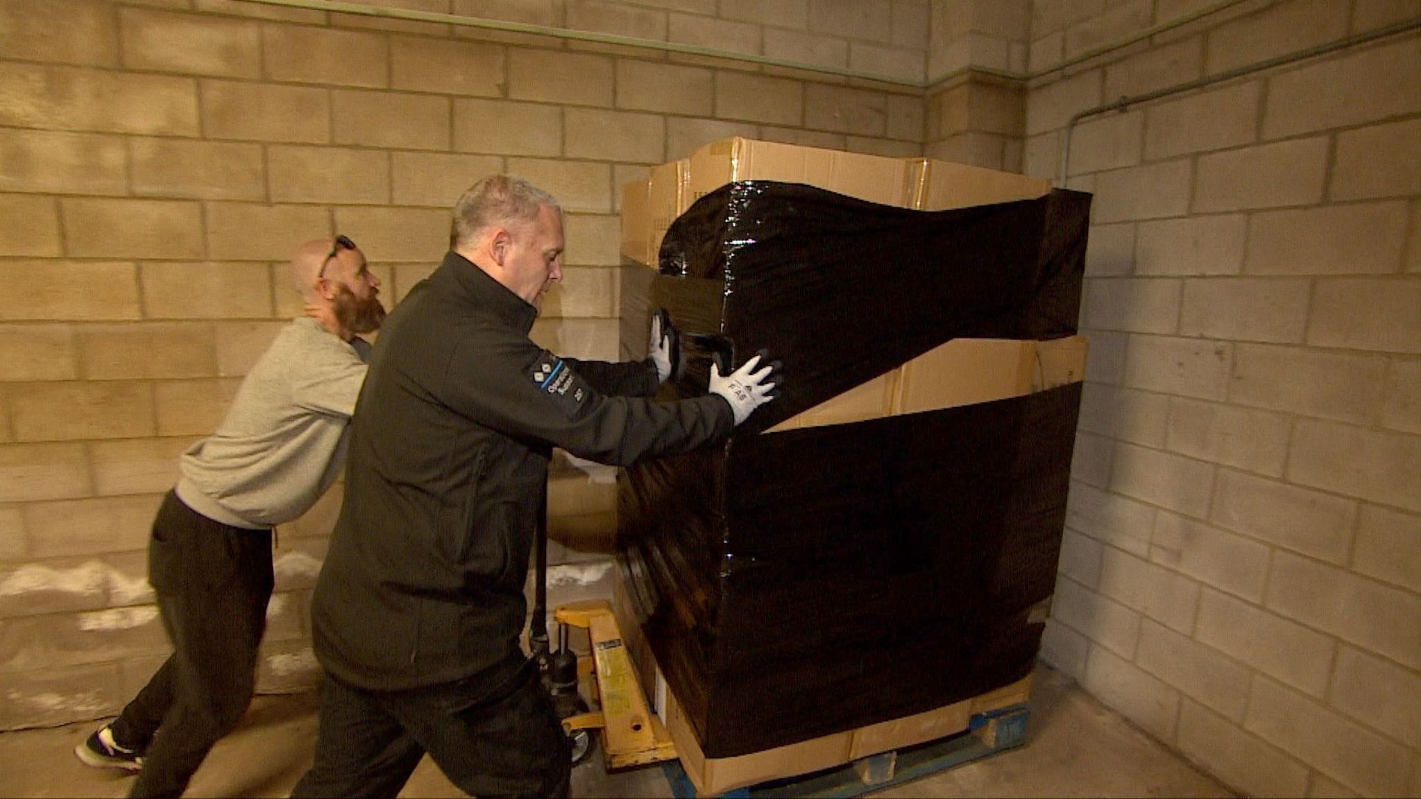 Two police officers pushing a pallet laden down with cardboard boxes into the corner of a breeze block-walled room. It is the only pallet visible. The boxes are around the same height as both officers and are partially wrapped in black cellophane. One officer is wearing a police jacket and we can see he is an inspector. The other is wearing a grey sweatshirt, dark joggers and casual shoes.  