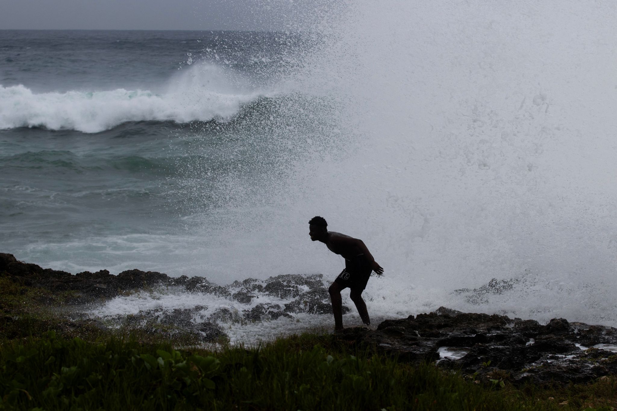 A man standing on the rocks is silhouetted against the waves and spray of the sea