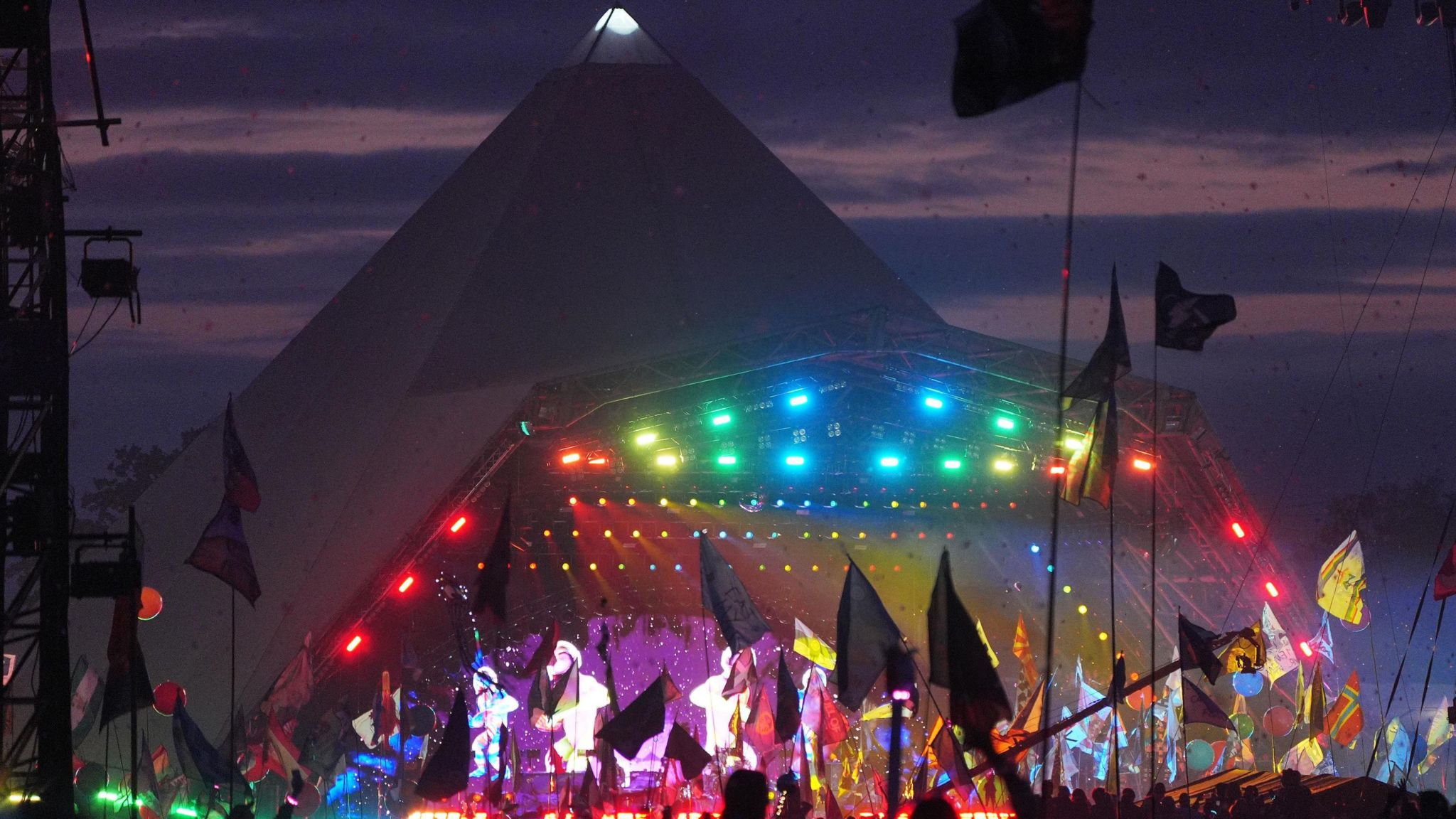 The Pyramid Stage at Glastonbury Festival during the Coldplay show. It is covered with a rainbow of lights as members of the crowd wave flags in front of it.