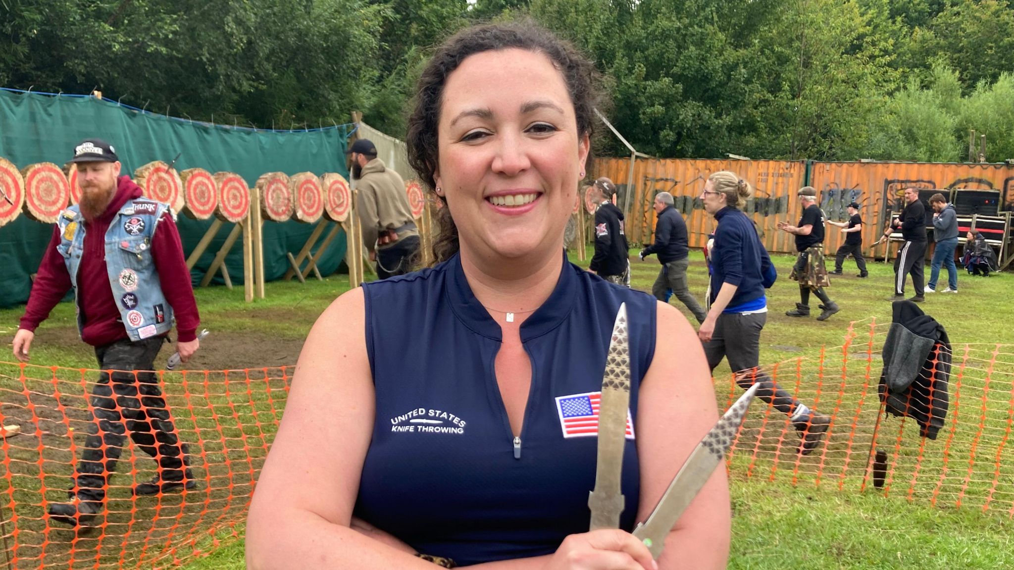 Natalie Murray wears a blue sleeveless top with a US flag logo and holds two metal knives. She stands in front of a grassy area where eight people throw knives towards round targets.