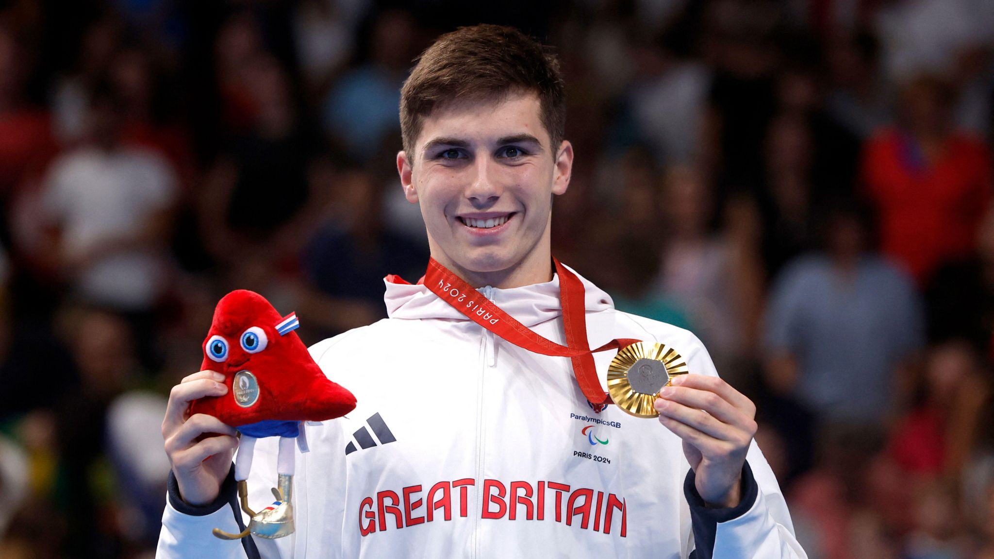 Will Ellard pictured in his white ParalympicsGB kit collecting his gold medal, following his 200m freestyle win at the Paralympics. He has short black hair and is smiling at the camera, clutching his medal in one hand and a toy Paralympics mascot in the other. 