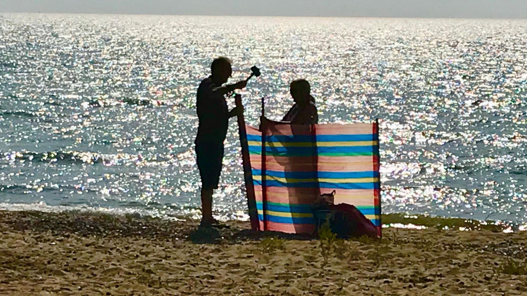 A man hammering the post of a windbreak into a beach, while a woman holds it and the sea sparkles behind them