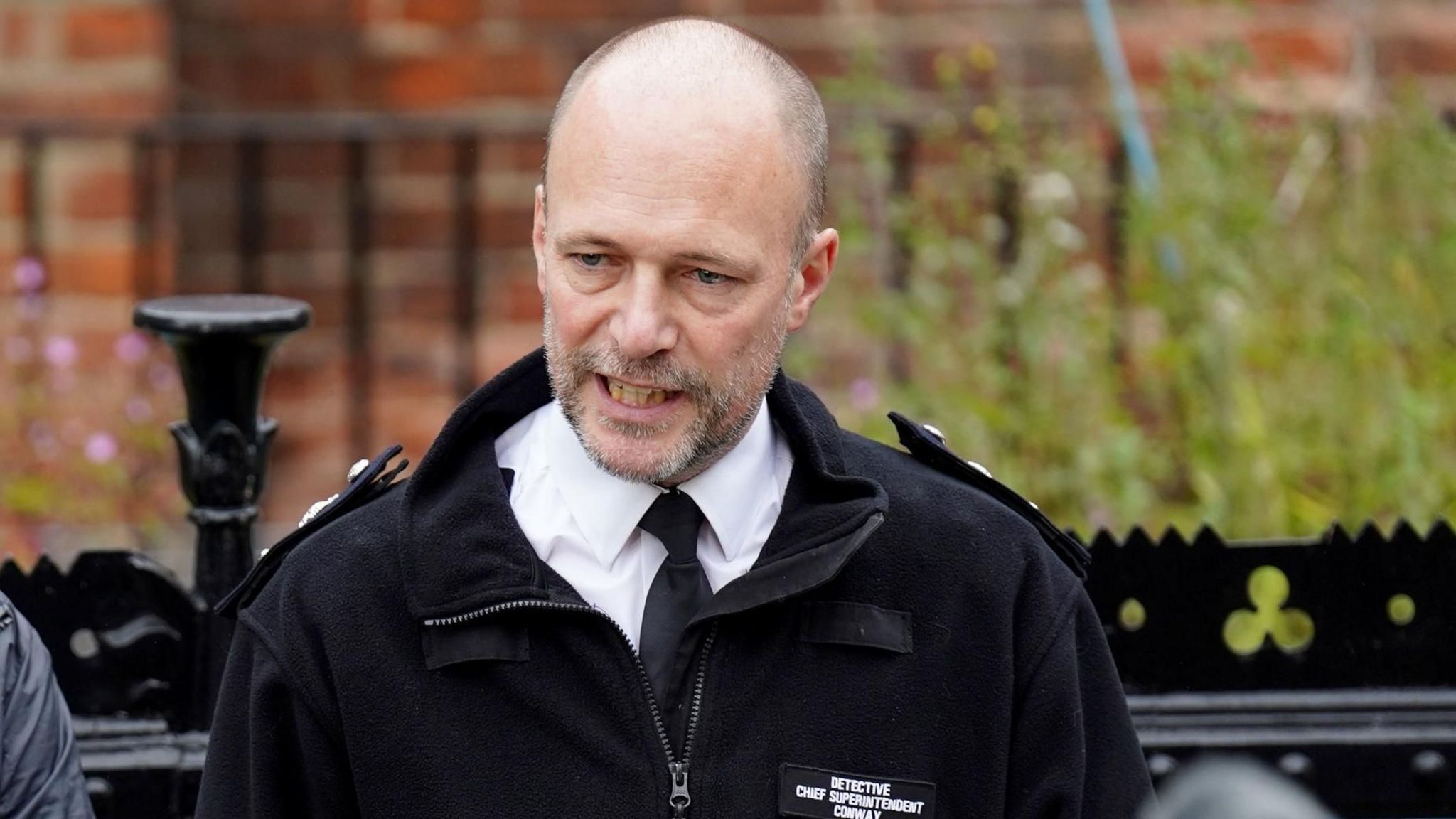 Det Ch Supt James Conway, from the Metropolitan Police. The police officer has a shaved head and short grey beard. He's pictured outside in front of a wall and iron fence, wearing his police uniform - a black jacket and tie over a white shirt. 