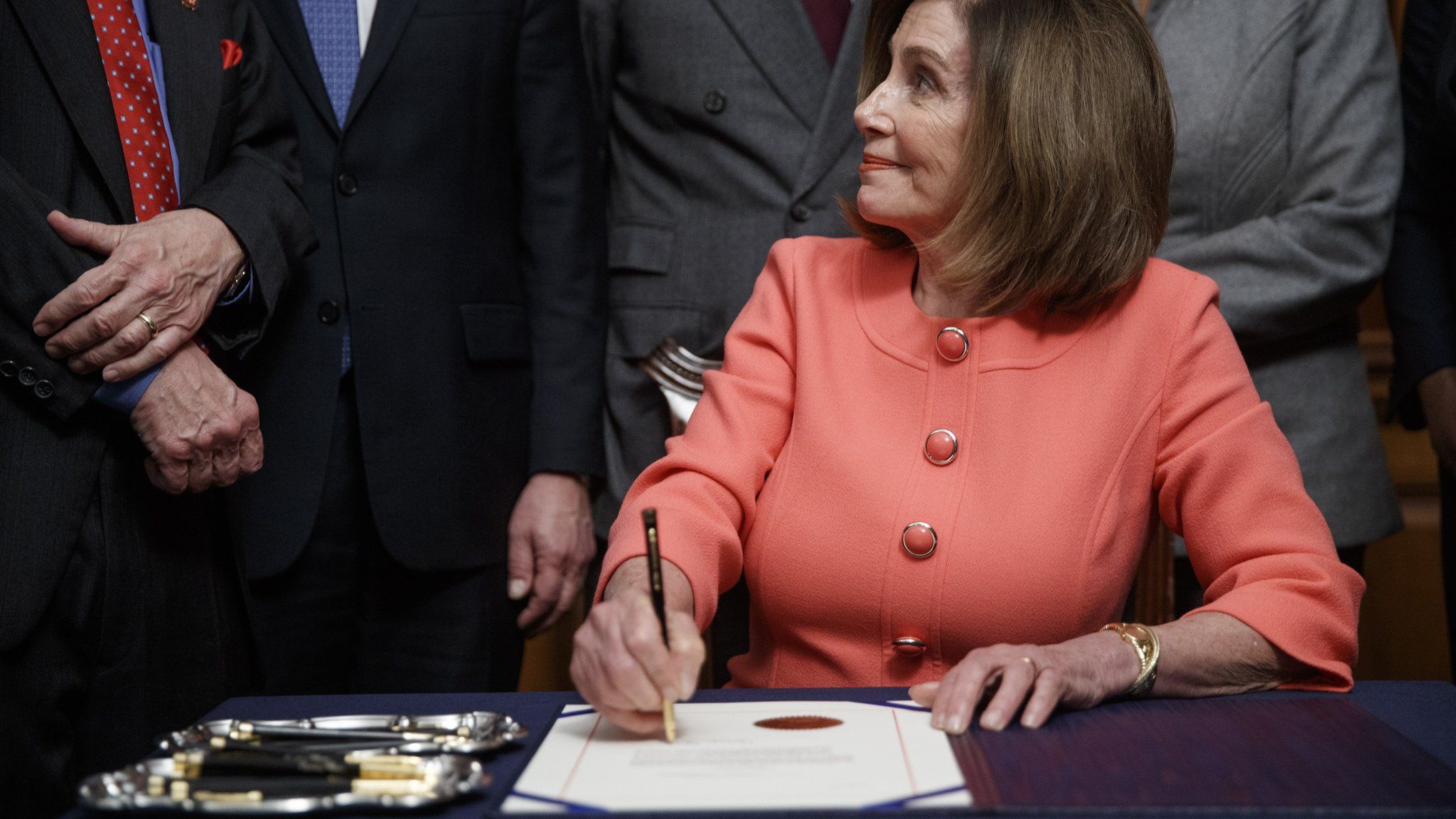 Nancy Pelosi signs two articles of impeachment, with two trays of pens by her side