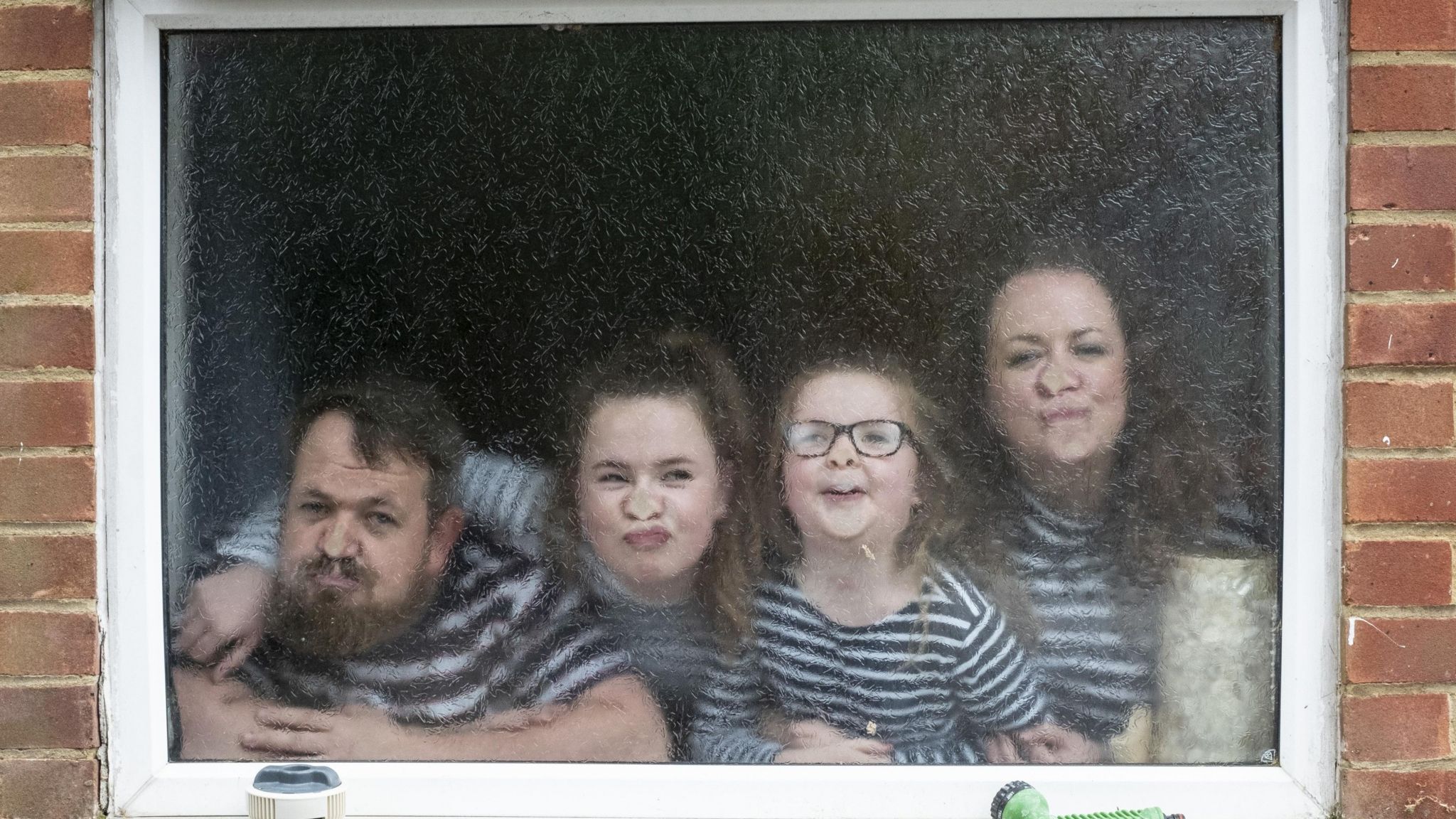 A family playfully are pressing  their noses up against a frosted window. From left to right there is a man, a girl, a younger girl and a woman. They are all wearing black and white striped tops. 