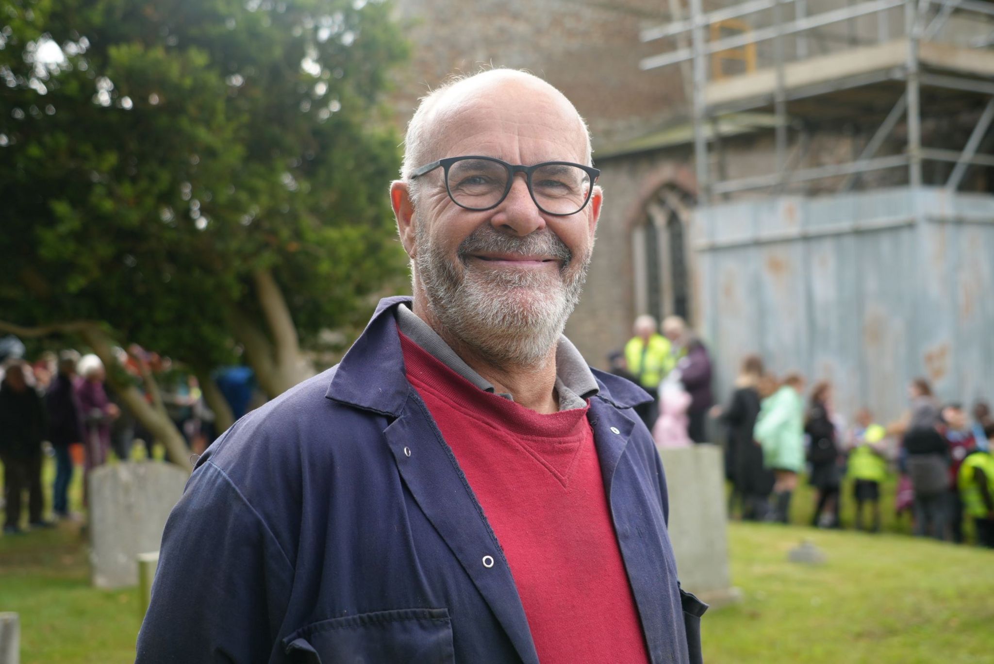 Bell installer Neil Thomas is smiling and wearing a red sweatshirt and blue jacket. There are many schoolchildren and adults behind him next to a church.