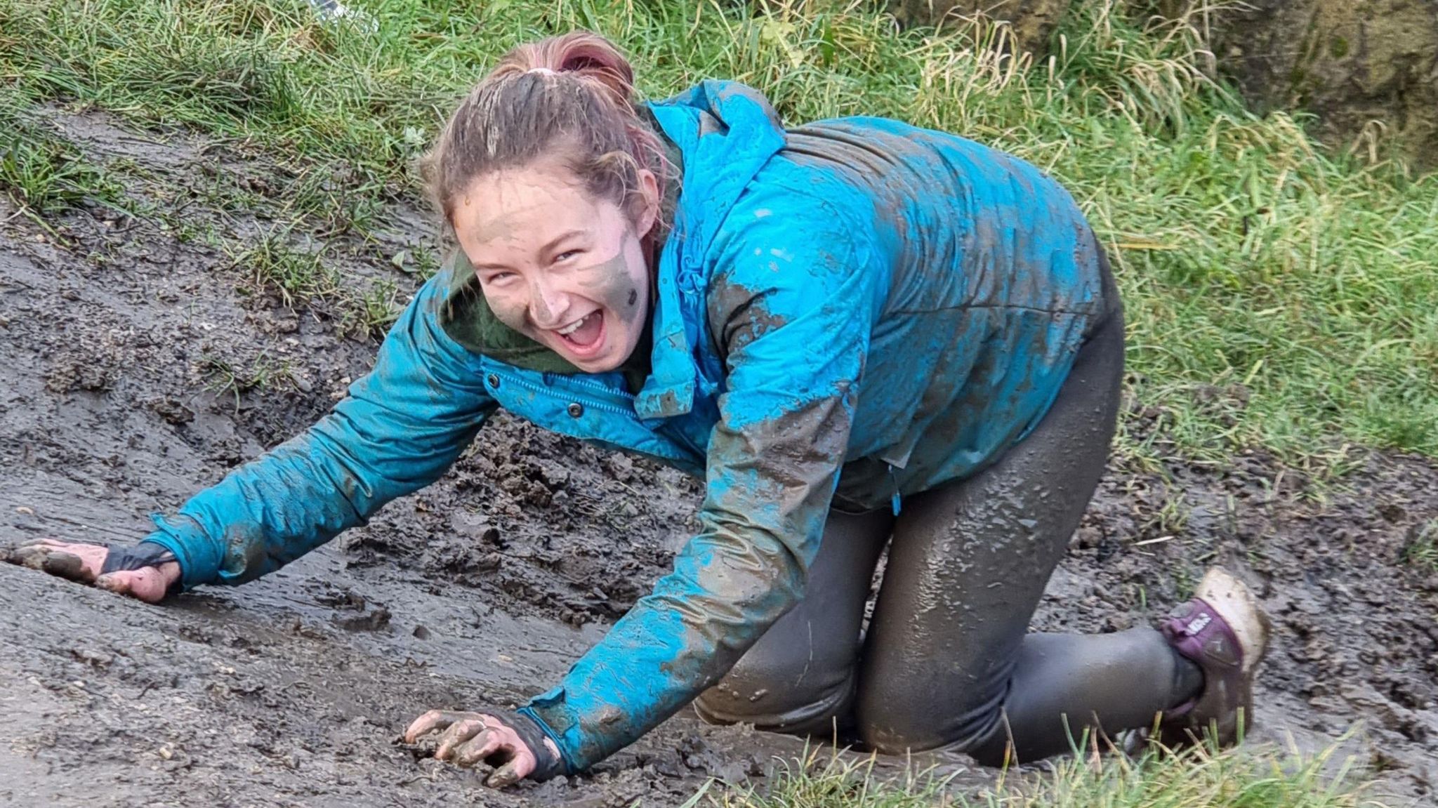 A white girl in a blue waterproof jacket kneels in the mud, smiling, mud on her face and in her hair 