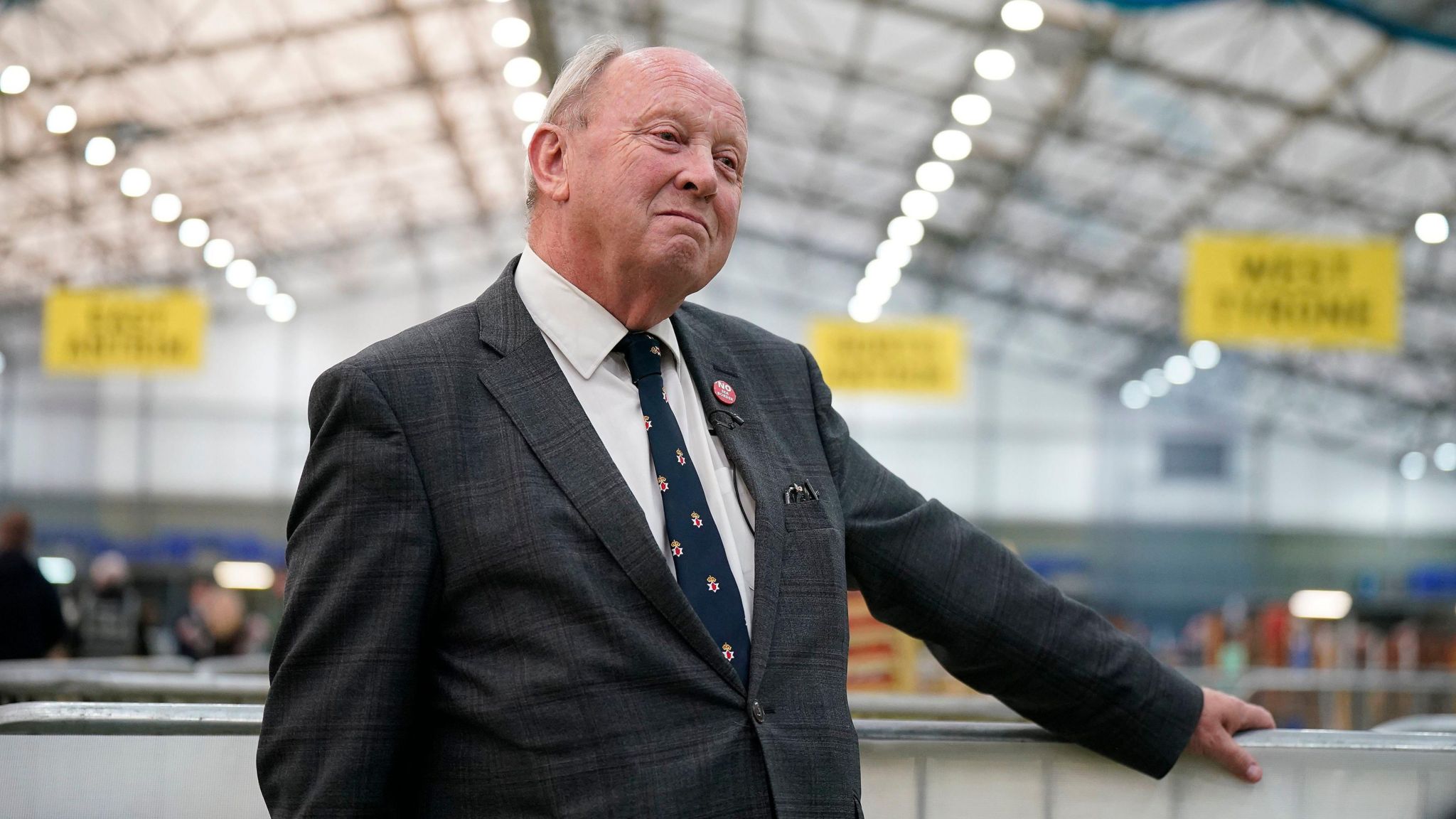 Jim Allister standing in the centre of the photo holding onto a railing with his left hand. The background is a blurred count centre. 