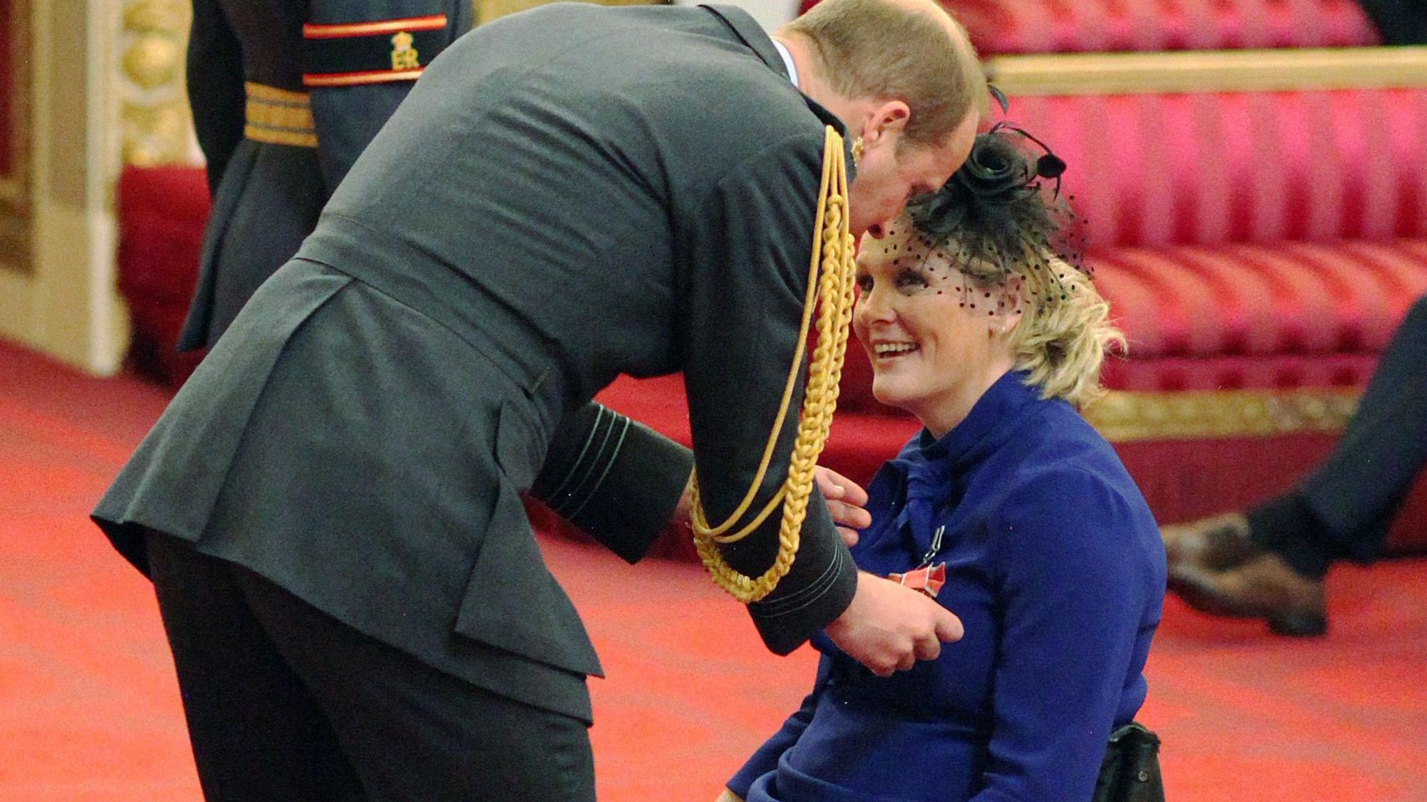 Ms Lomas being made an MBE by the Duke of Cambridge at Buckingham Palace in 2017. He, in a dark military suit with gold braiding on the shoulder leans down to pin the honour on her purple top.