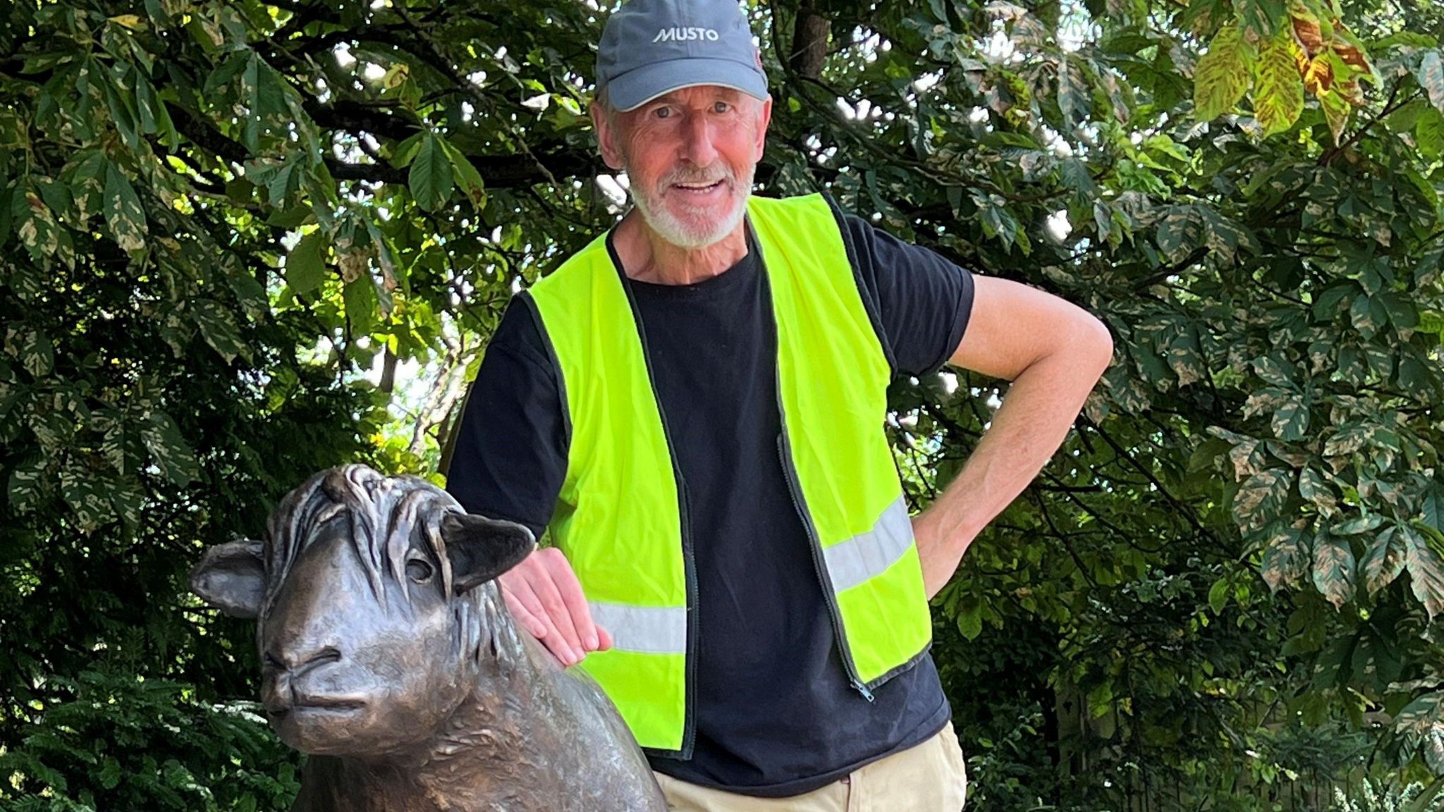 Jonathan Sanders smiles at the camera wearing a high vis jacket and a cap, resting an arm on the bronze sheep statue