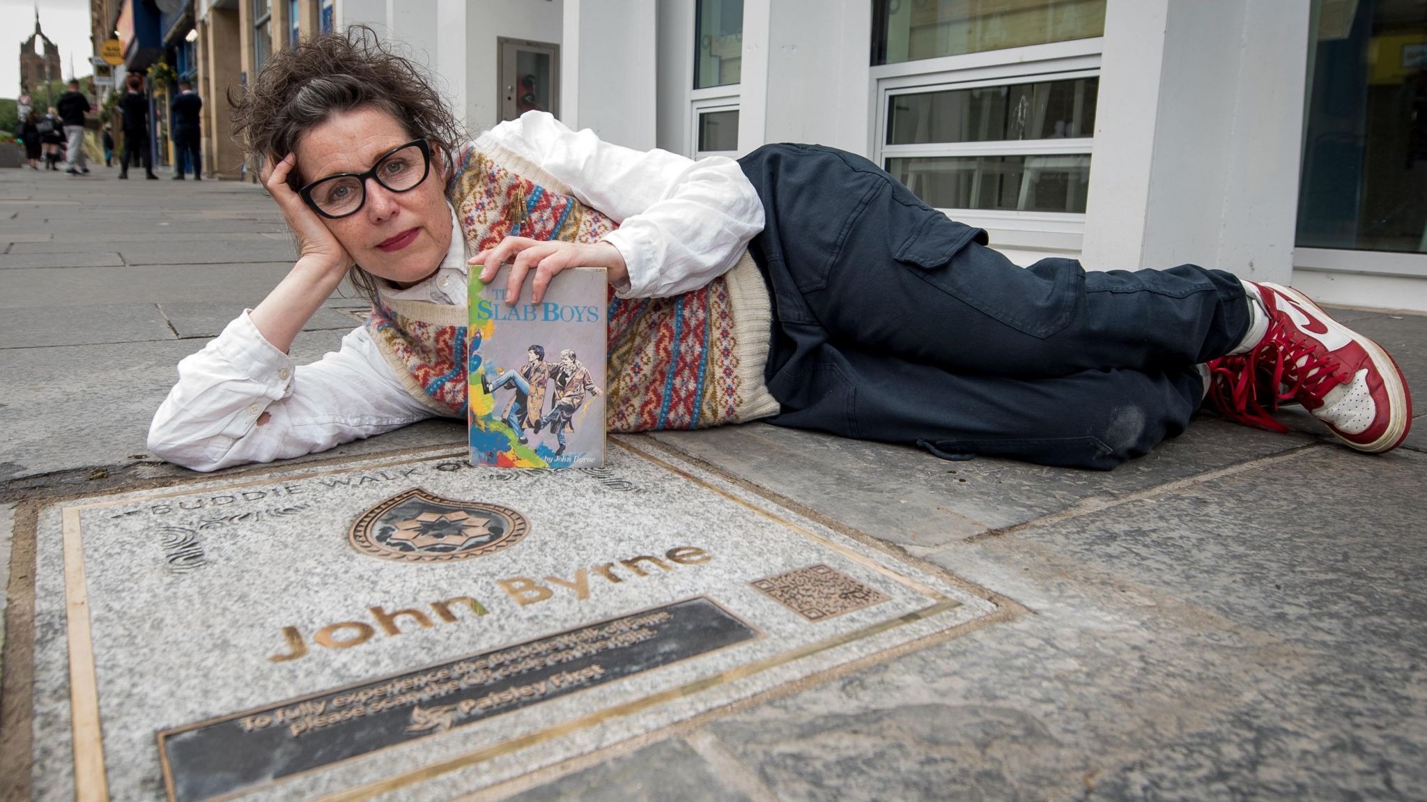 Celie Byrne lying on the ground beside John Byrne's walk of fame plaque. She's holding Byrne's Slab boys books
