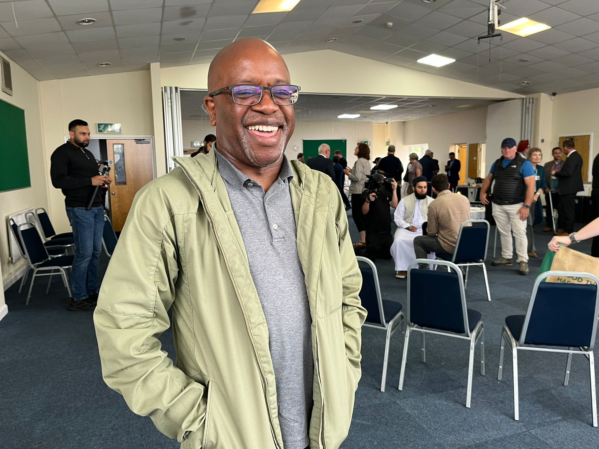 Reverend Geoffrey Eze wearing a light jacket and grey polo shirt, standing in a room full of people. 