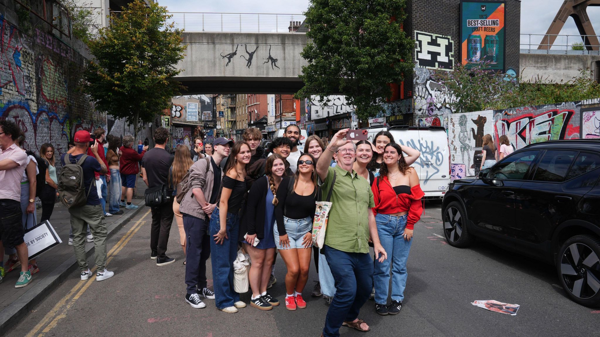 A group of people take a selfie on Brick Lane with the Banksy work in the background