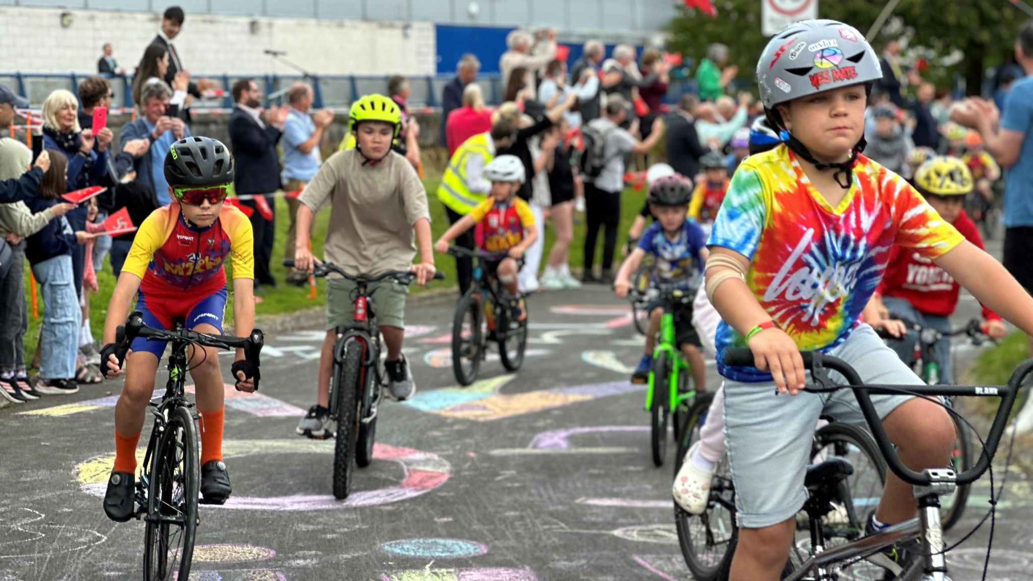 Children in colourful clothing and bicycle helmets riding on the raceway with crowds waving flags in the background.