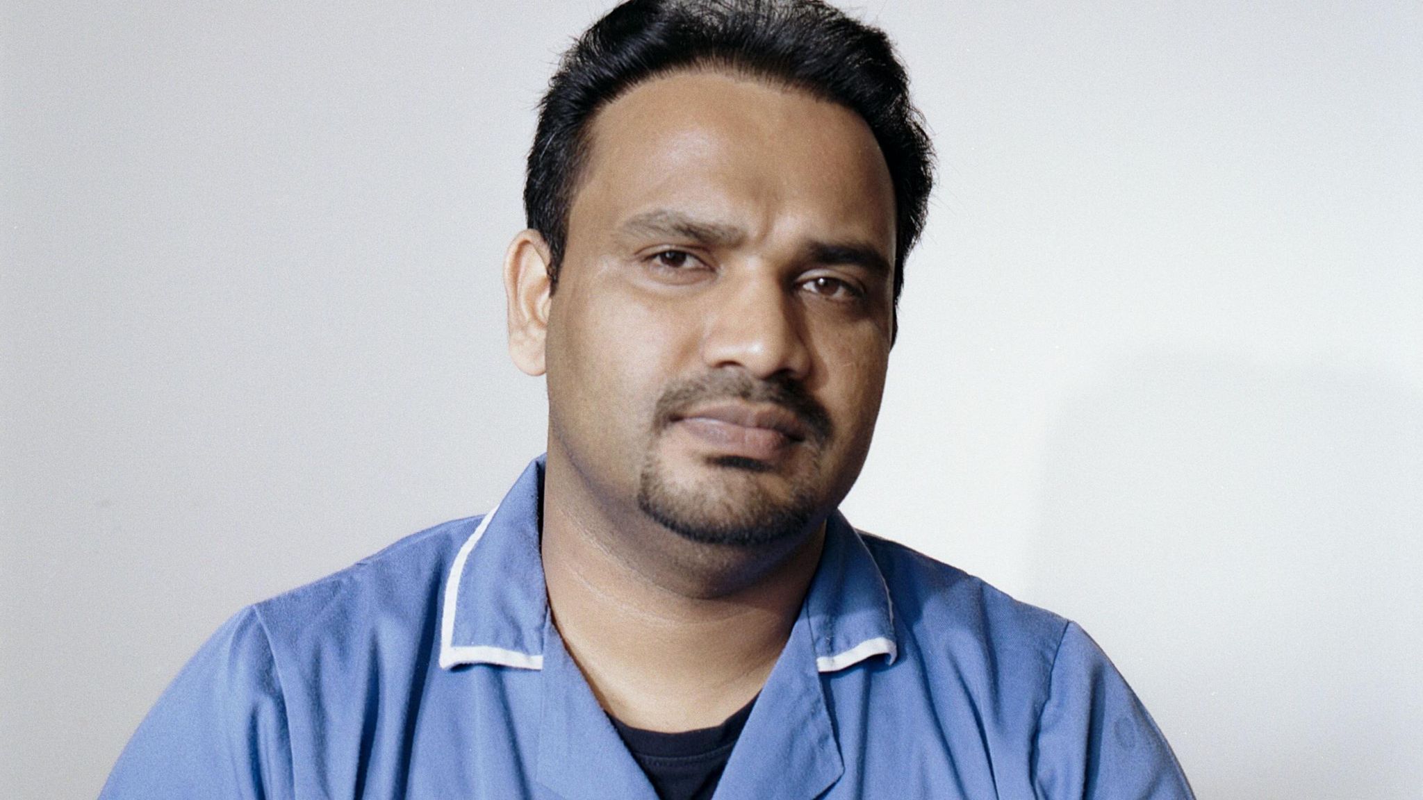 Portrait of Nidheesh Madhava. He has short dark hair and facial hair. He is wearing a blue care uniform with a collar. He is looking directly at the camera with a slight smile. 