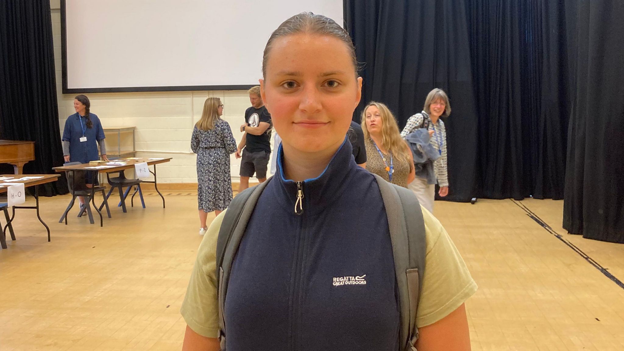 Kingsbridge Community College pupil Anna Petrenko looking at the camera in a sports hall after picking up her A-level results. She is wearing a navy blue gilet jacket and a light green t-shirt and has a back pack on