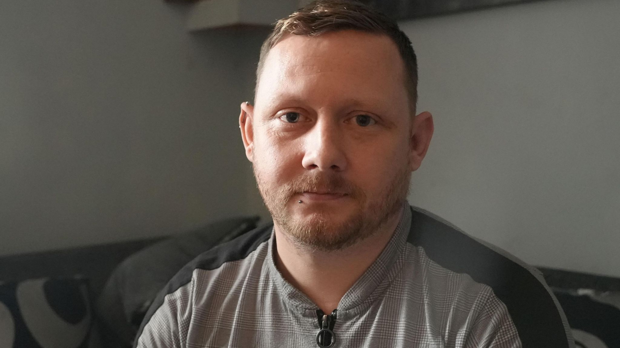 Gaz has short dark hair and a short beard. He is wearing a grey zip up top and is sitting on a sofa, looking into the camera.
