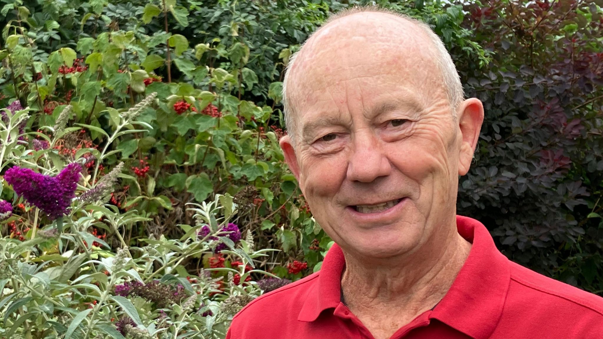 Resident wearing a red shirt and standing in front of a buddleia bush with purple blooms