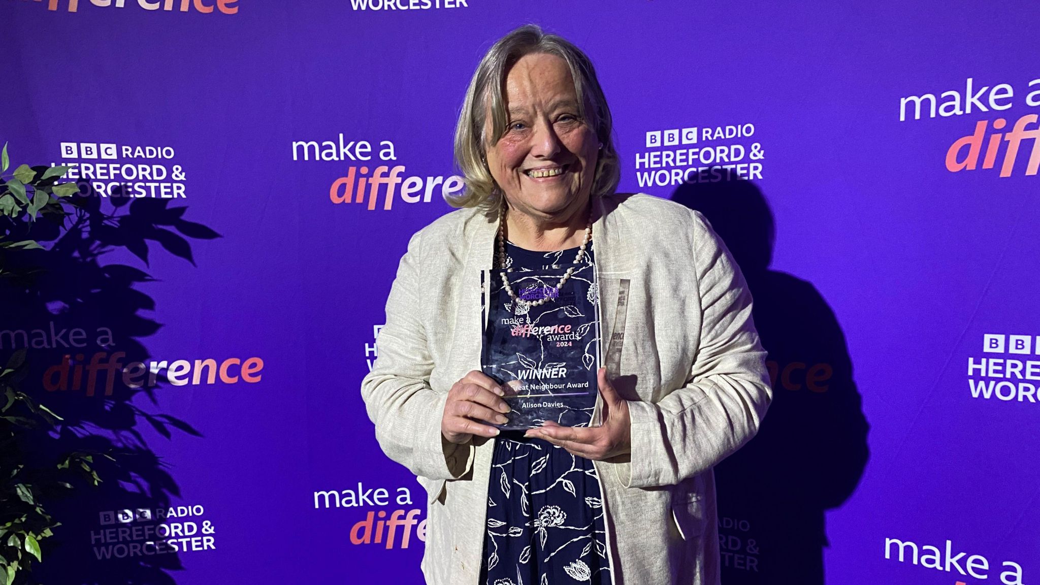 A woman stands in front of a BBC backdrop. She has blonde hair and is wearing a white jacket with a blue and white top. She is holding an award