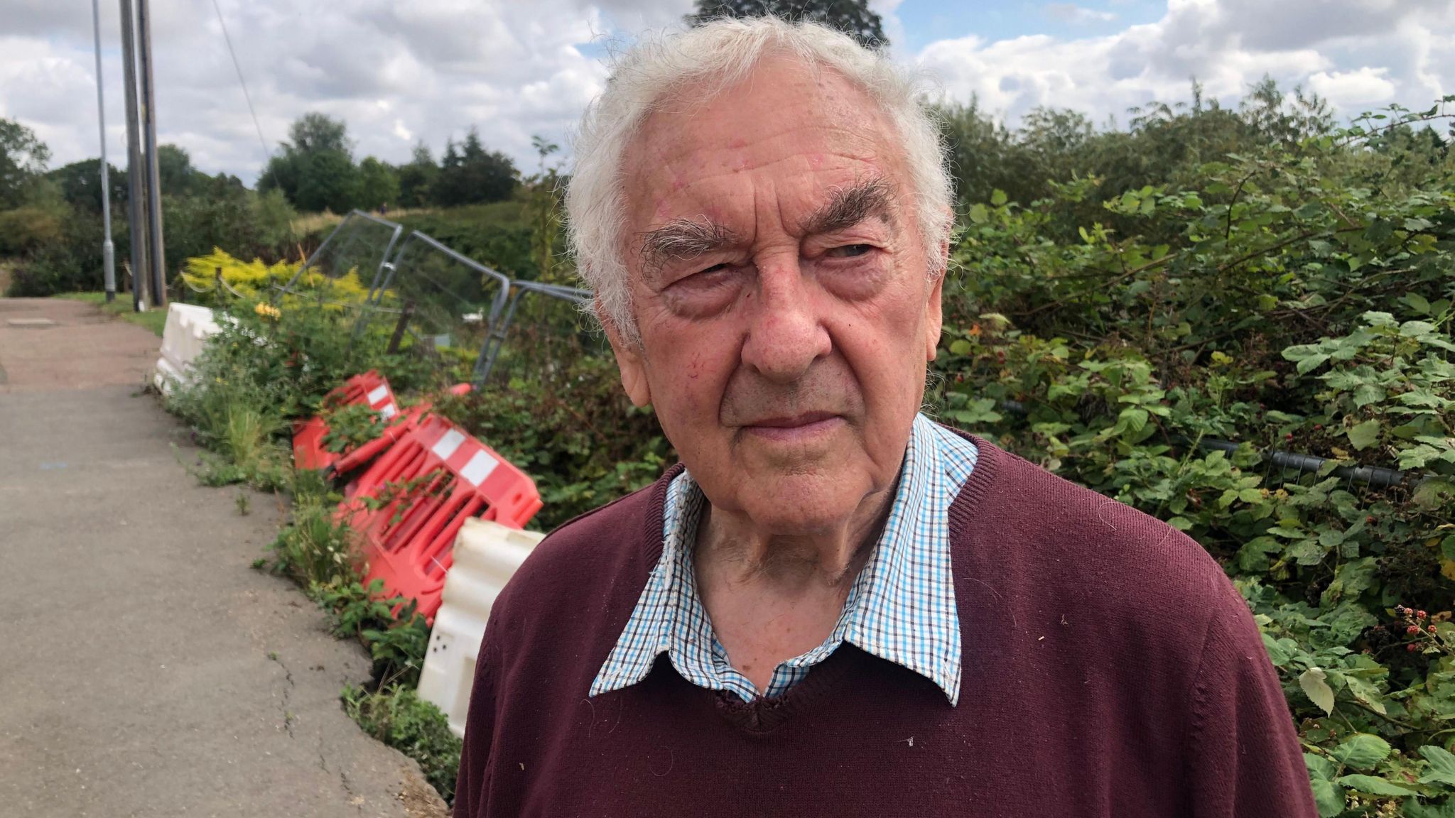 Bill Hill looks to the right of the camera as he stands on the pathway. Behind him is red safety fencing and silver fencing on its side leaning against green shrubbery. He is wearing a burgundy jumper and chequered shirt. 