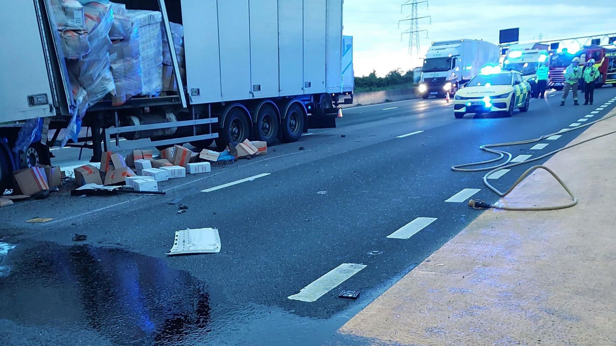 Emergency service vehicles are at the scene, the large white lorry to the left has the side broken open and boxes are on the floor and spilling from the side
