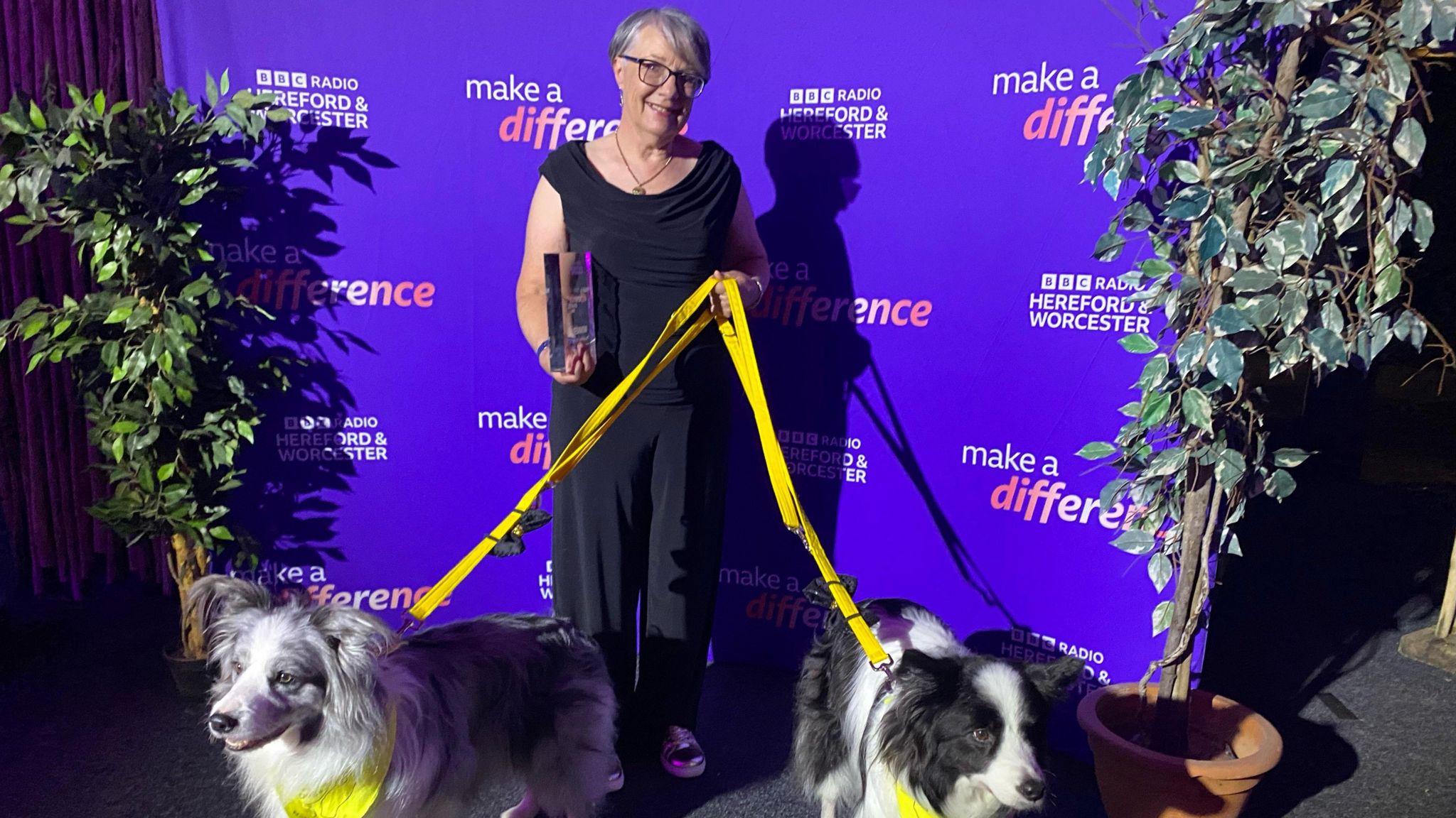 A woman standing in front of a BBC backdrop. She is holding an award and has two therapy dogs on a yellow lead