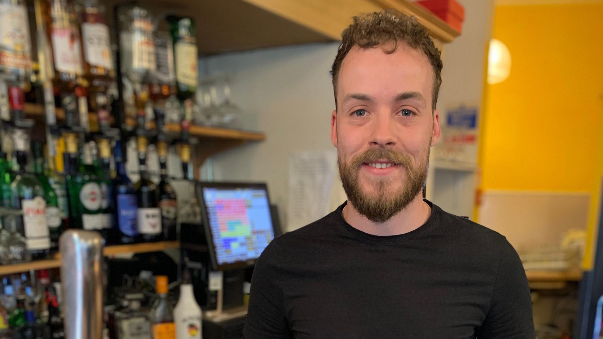 George Sperrin is behind the bar of a pub with a till machine behind him along with numerous alcohol bottles. He has a beard and curly dark hair and wears a black T-shirt