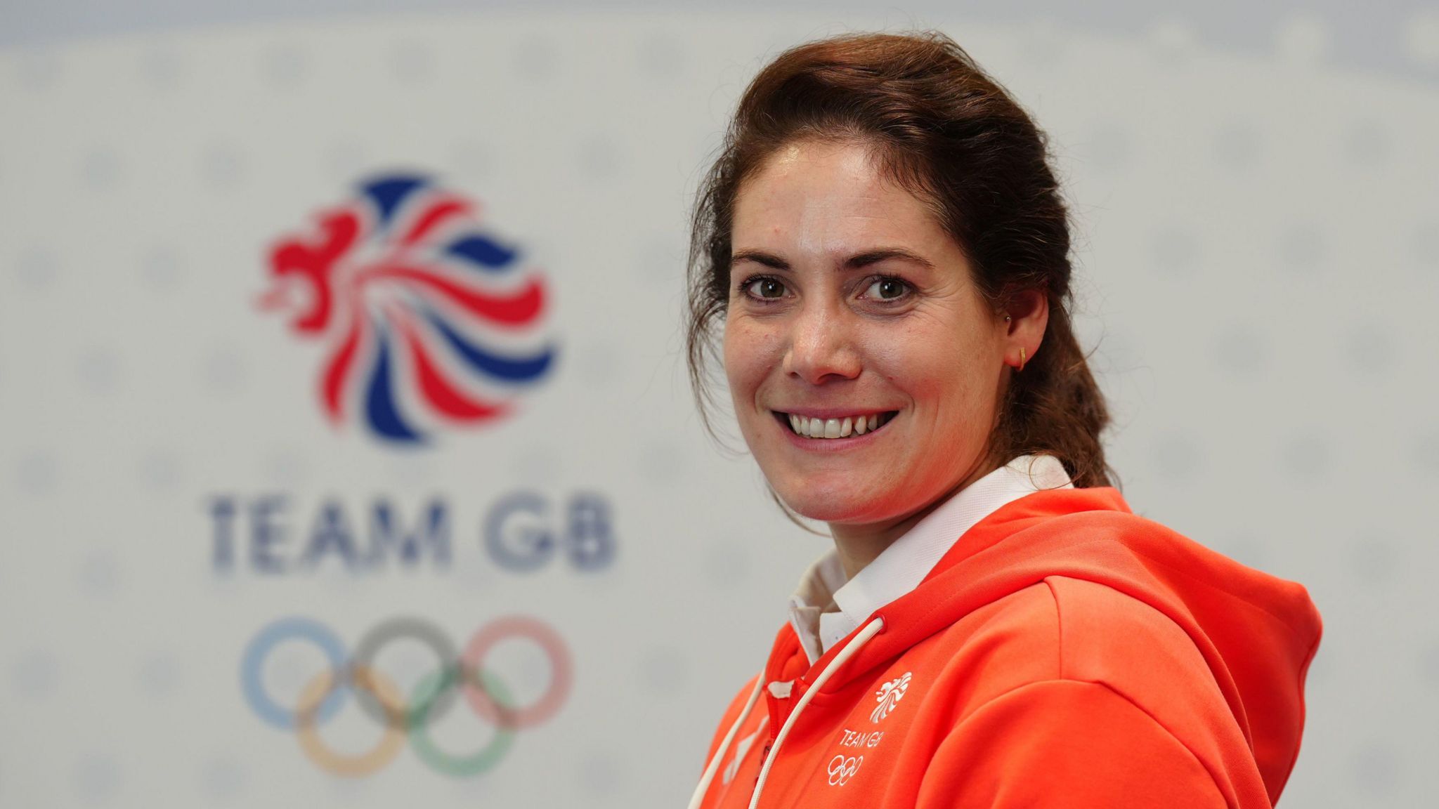 Kate French wearing a neon orange Team GB hoodie with the Team GB and Olympic ring logos in the background. She is turned to the side, smiling at the camera and her brown hair is up in a ponytail.