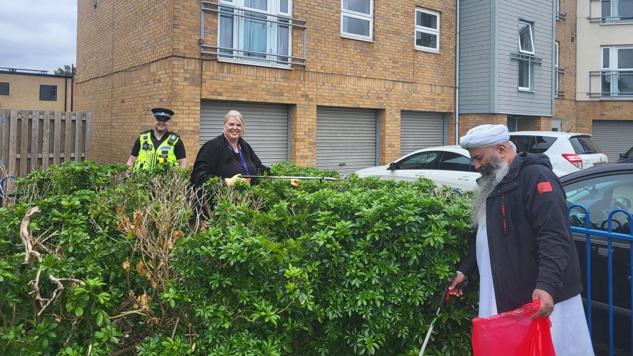 Two people and police officer picking up litter around some bushes