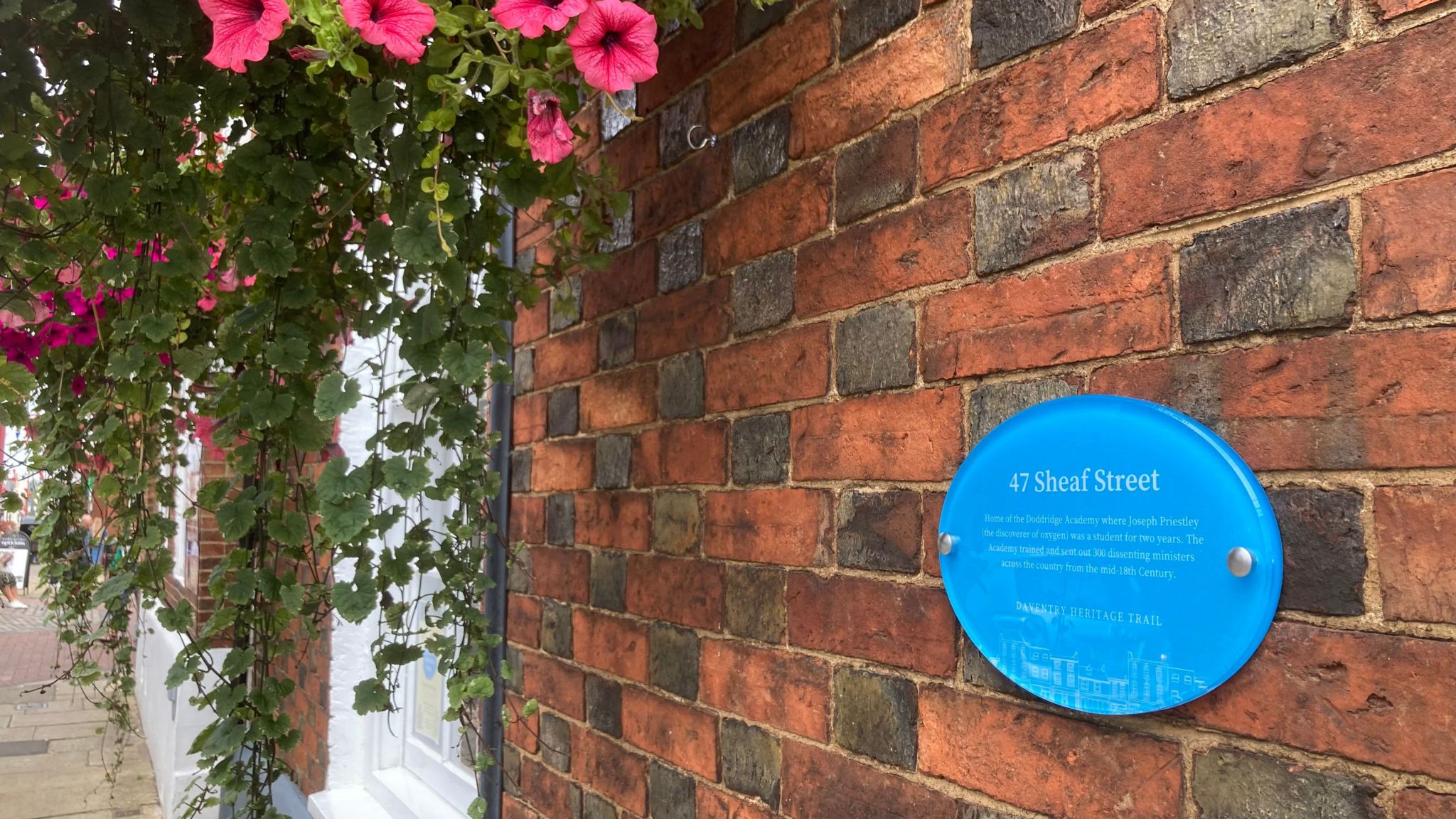 Brick wall bearing a blue plaque headed "47 Sheaf Street", alongside a hanging basket with pink flowers