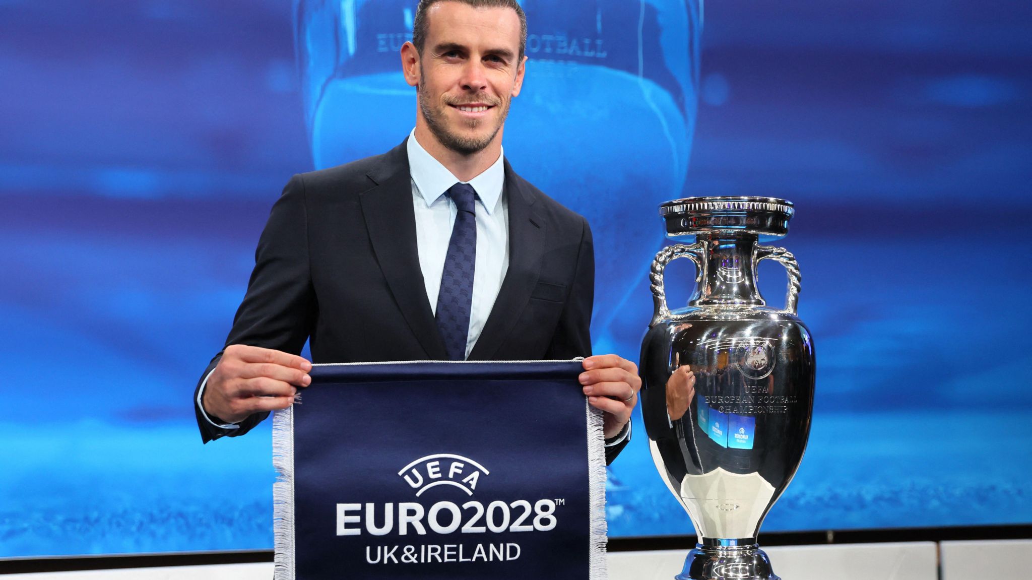 Former Wales footballer Gareth Bale holds up a blue pennant with the words Uefa Euro 2028 UK and Ireland