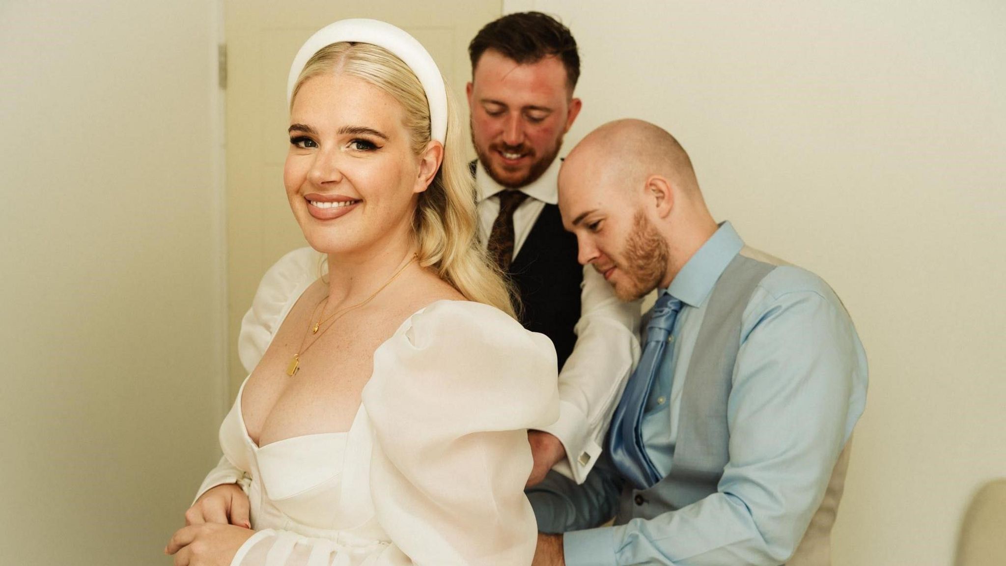 Brooke Jones is standing in a white wedding dress, wearing a white headband. Behind her is a bald man in a blue suit, adjusting the back of the dress. Next to him, is a man with brown hair in a suit