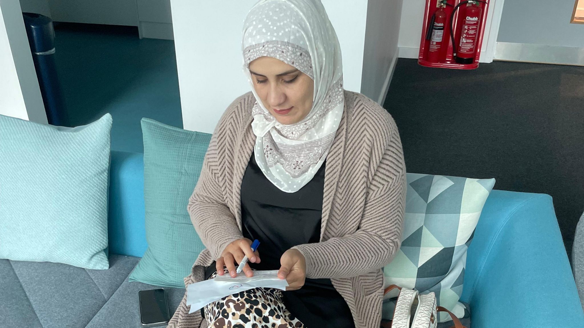 A young woman wearing a hijab, sitting on a sofa and working with a pen and paper