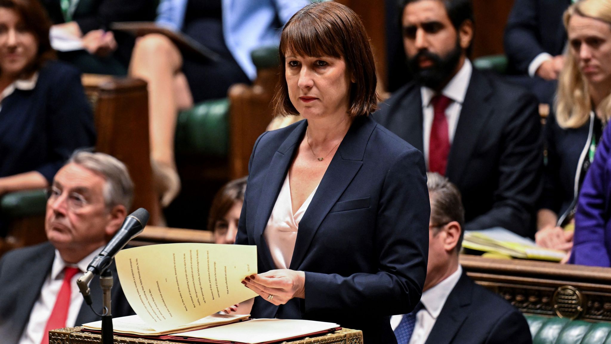 Chancellor Rachel Reeves at the dispatch box addressing the House of Commons. She is holding paperwork and is standing, surrounded by seated MPs. 