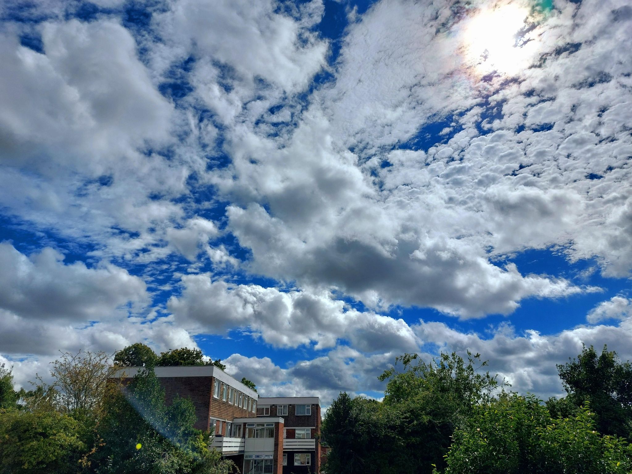 Clouds sprawled across bright blue sky with a red brick building and greenery below