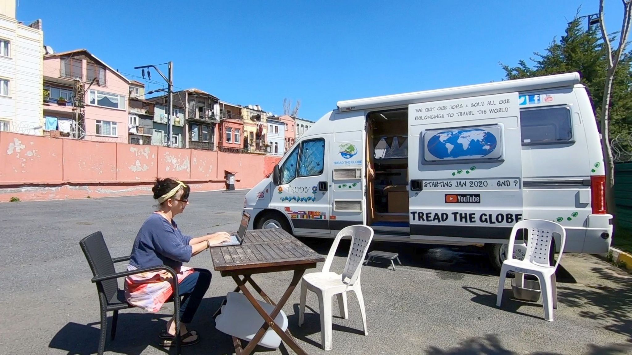 A woman sits using a laptop, which is resting on a wooden table. A white camper van is in the background as well as some plastic chairs. Further in the distance is a row of housing/apartment blocks