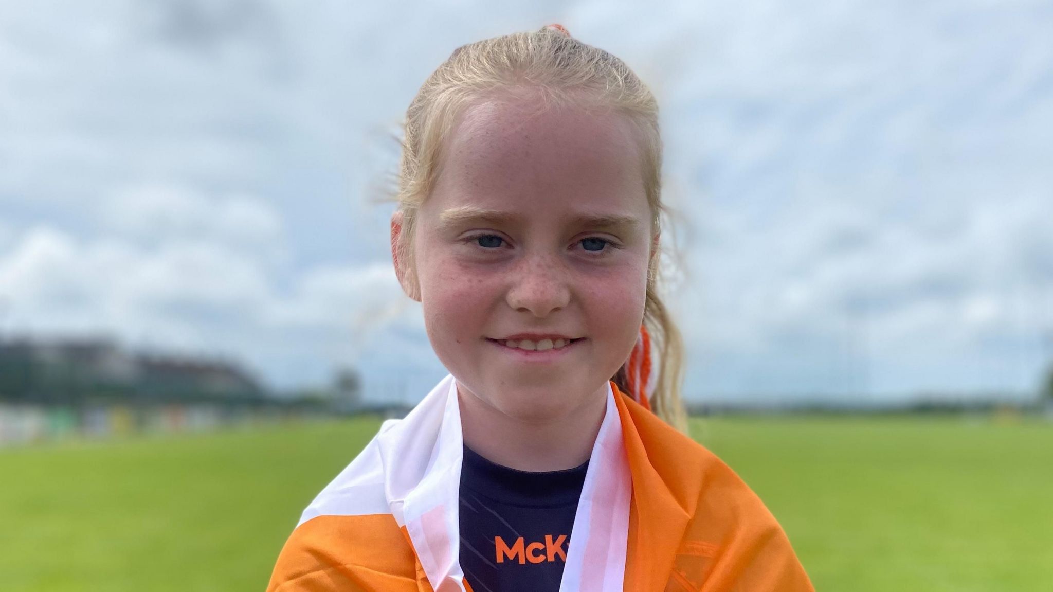 Sarah is smiling into the camera. she has her blonde hair tied into a ponytail and has an orange and white flag draped around her shoulders.