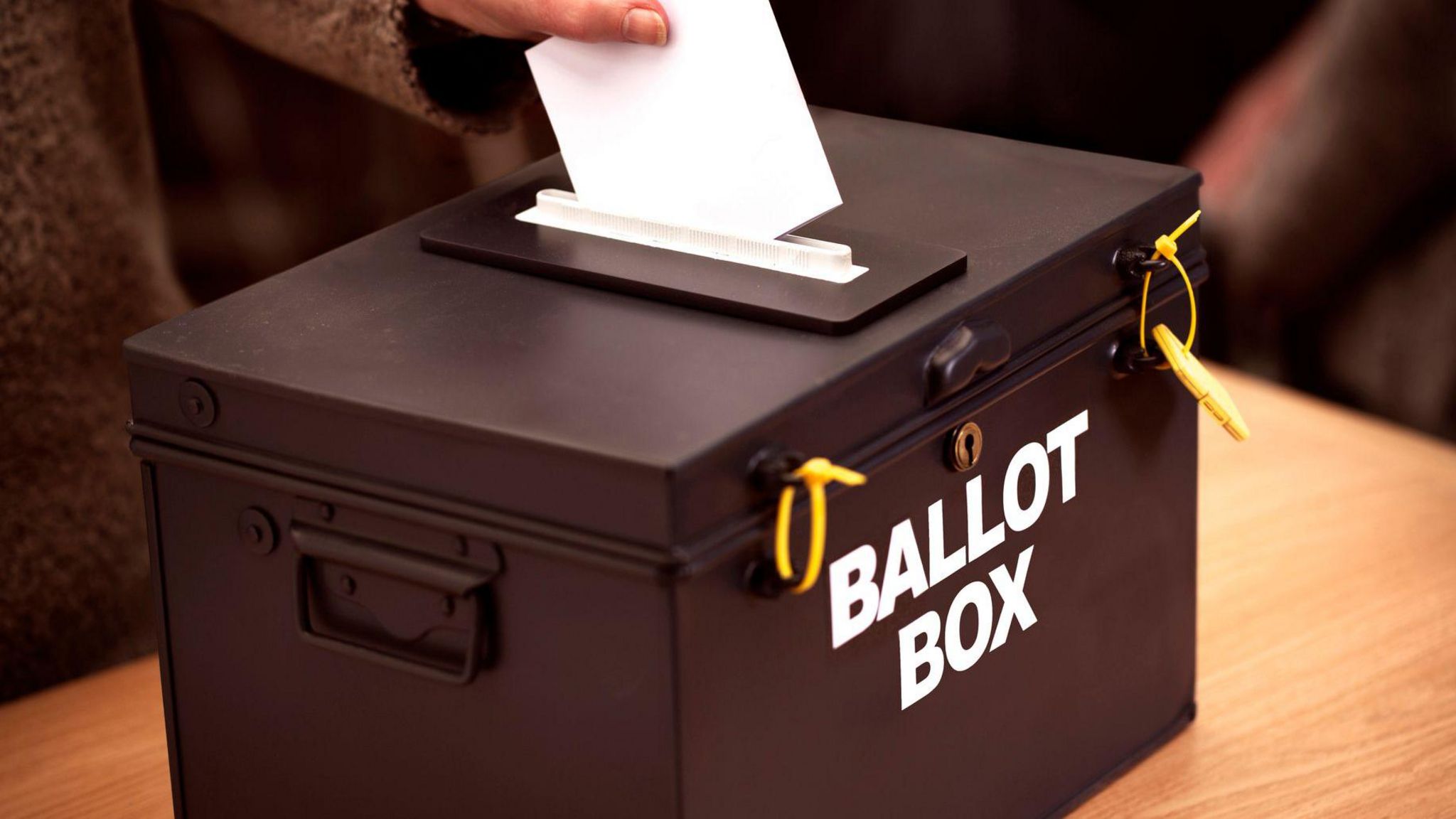A black box which reads Ballot Box is on a table, someone is posting a ballot paper through the hole in the top of it