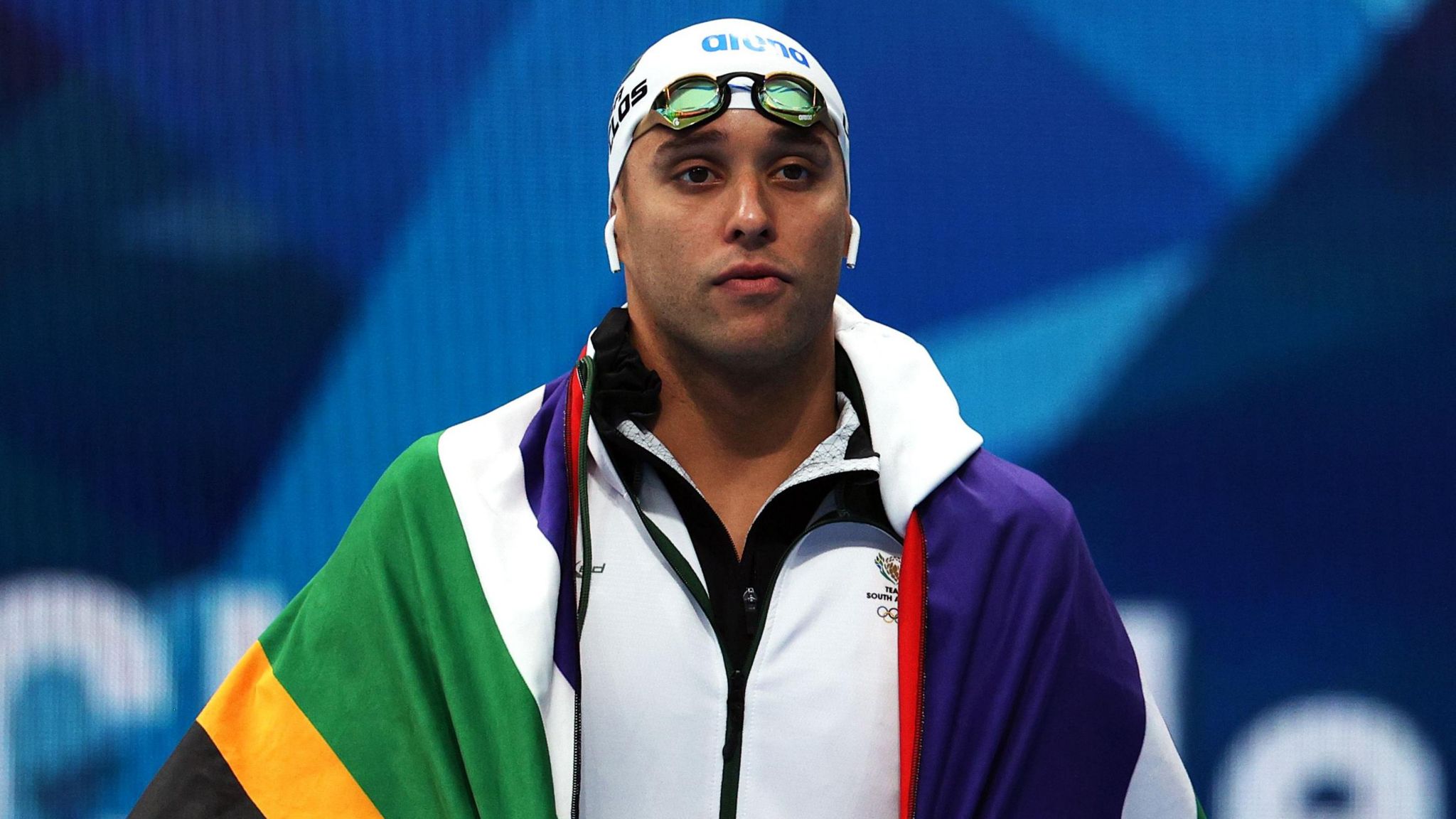 Chad le Clos, draped in a South African flag and wearing headphones, is pictured with a blank expression before a swimming race