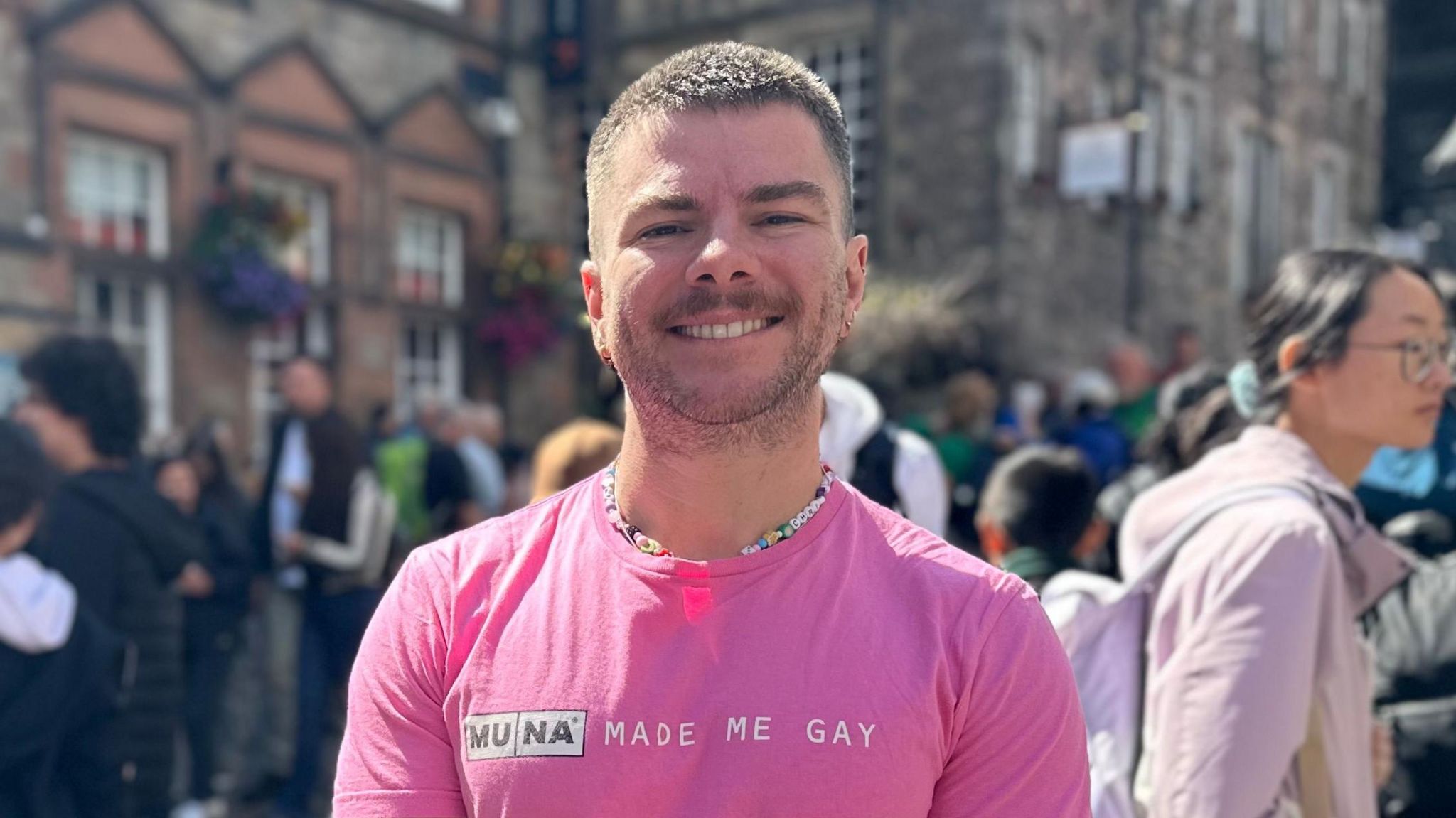 Chris Hall. Chris is a white man in his 20s with cropped brown hair and a stubbled beard. He smiles at the camera while standing on a busy street in Edinburgh for the Festival Fringe. He wears a bright pink T-shirt and a beaded necklace. 