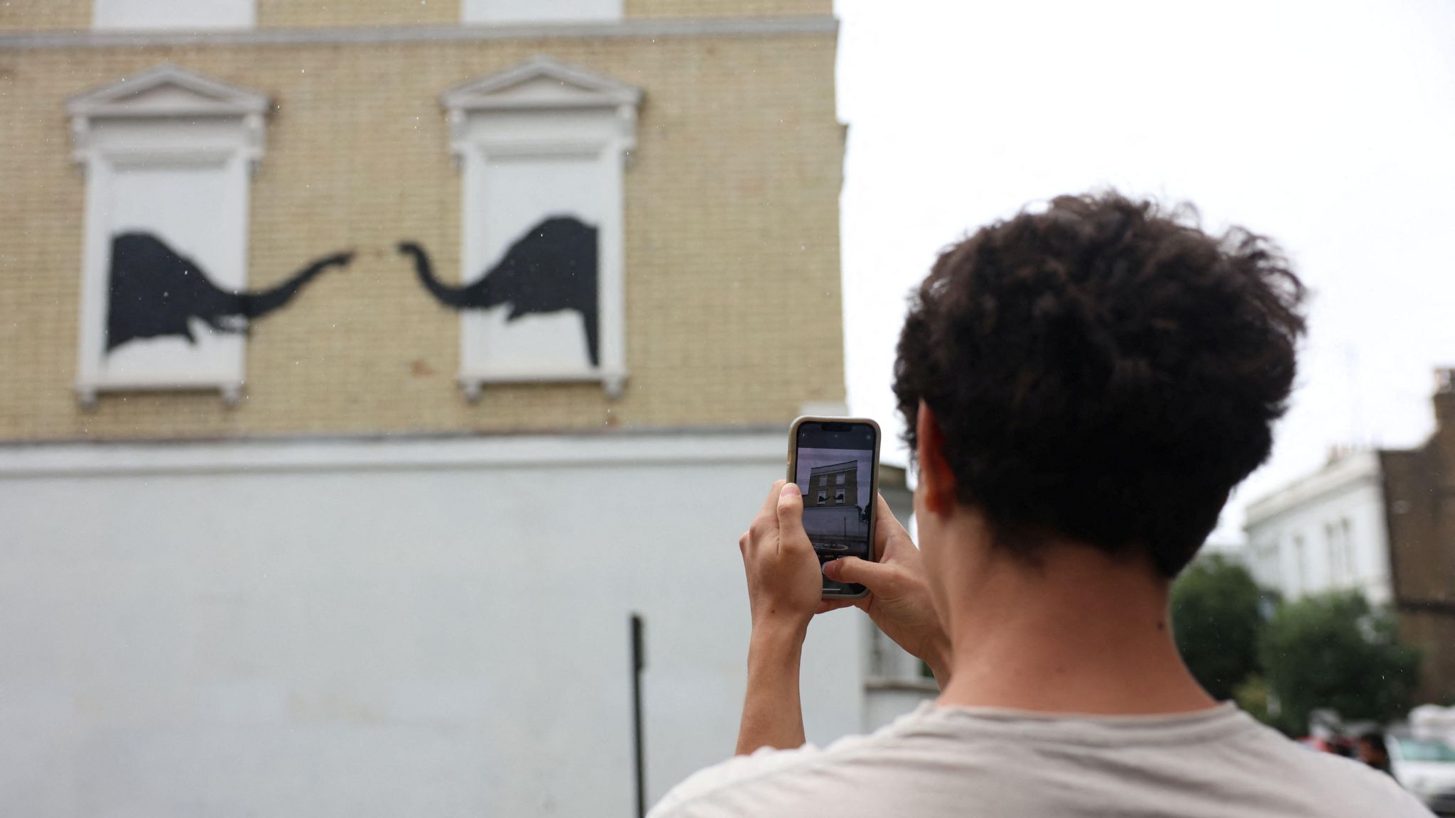 A man takes a picture of the Banksy work of two elephants with his phone