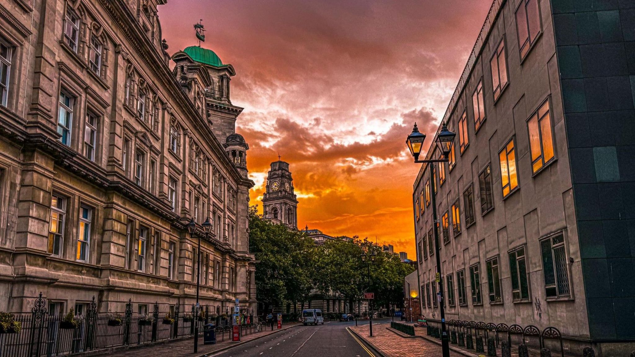 An orange sunrise over a city street with buildings on either side, a tower and some trees. 