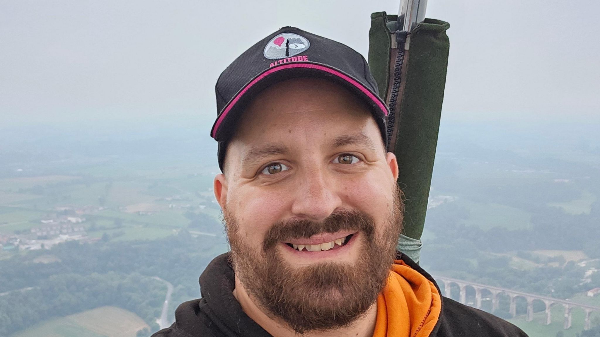 Jon London smiling and looking directly at the camera from a hot air balloon, with green fields and houses in the distance in the background